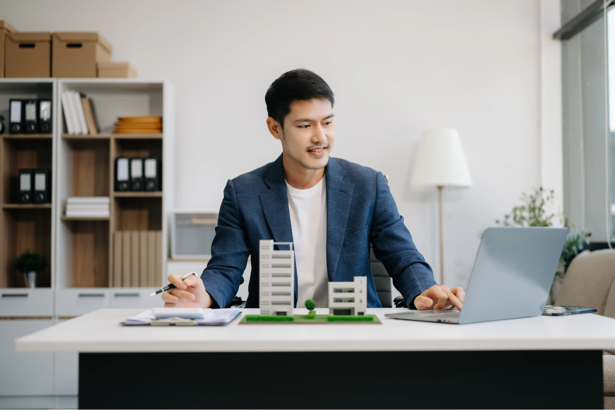 A man in a blue blazer sits at a desk, looking at a laptop. Surrounded by building models and documents, he appears to be working on a design project while considering business vs personal bank account​ options in his modern office.