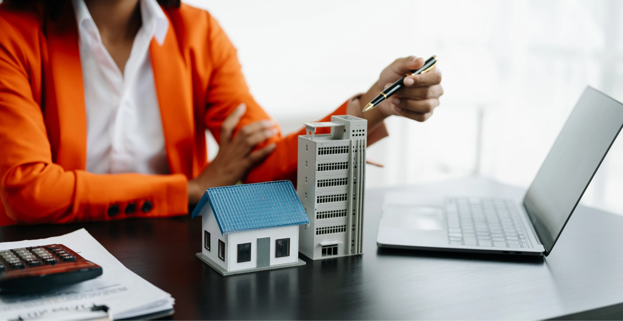 A person in an orange blazer gestures with a pen toward model buildings on a desk—next to a laptop, calculator, and documents—prompting a discussion about business vs personal bank account choices in real estate or property matters.