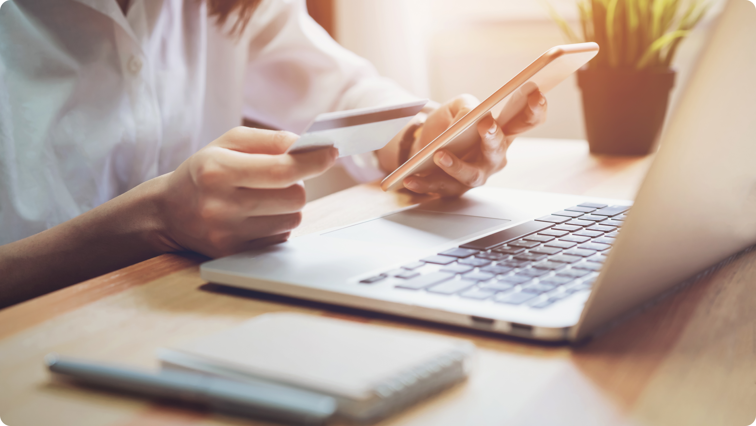 A person holds a credit card in one hand and a smartphone in the other, sitting at a desk with an open laptop, a notebook, and a pen in soft natural light.