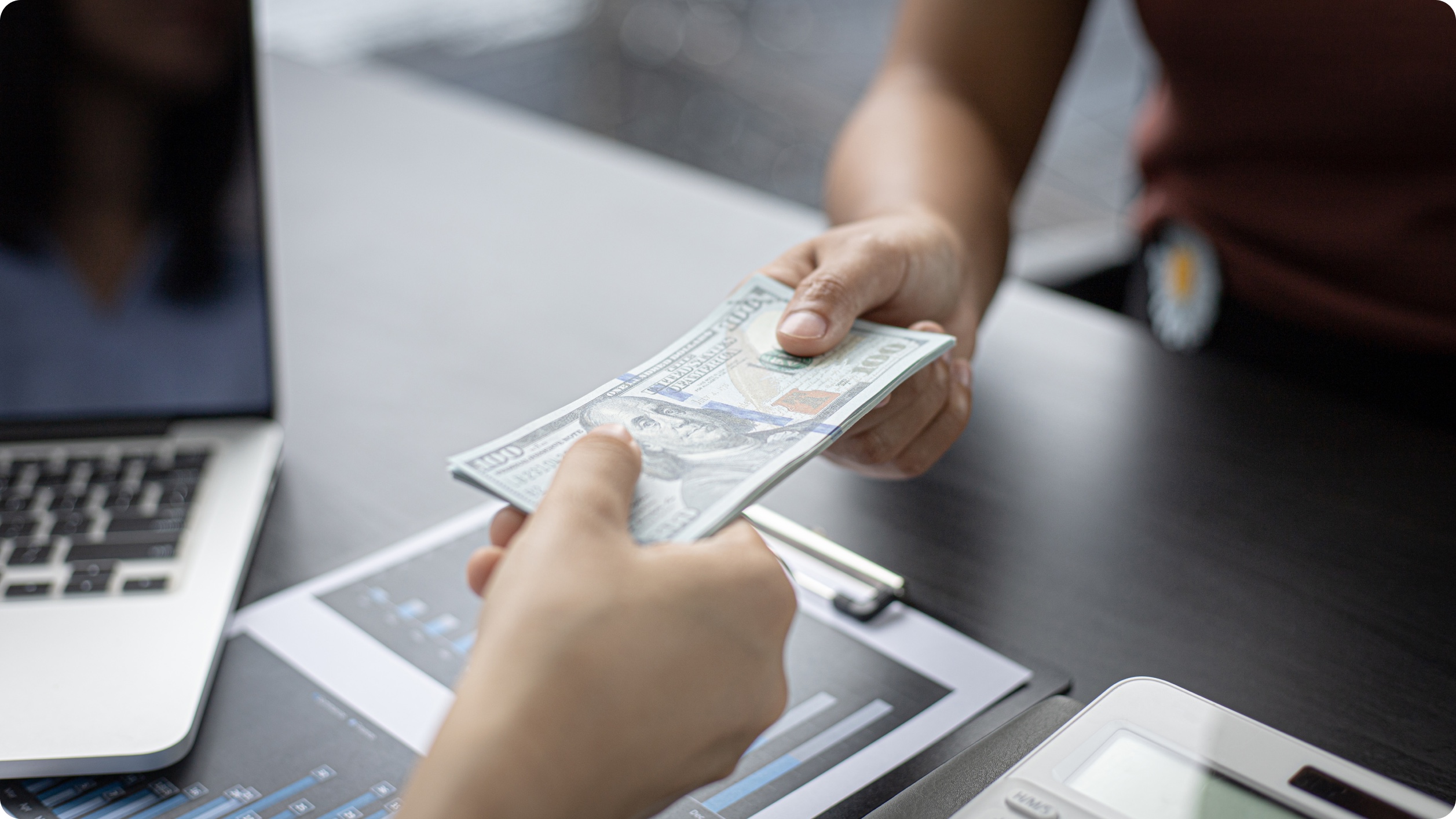 A person hands a stack of U.S. $100 bills to another individual across a desk with a laptop, a tablet displaying a chart, and a calculator—perhaps discussing how to choose a bank for managing their finances.