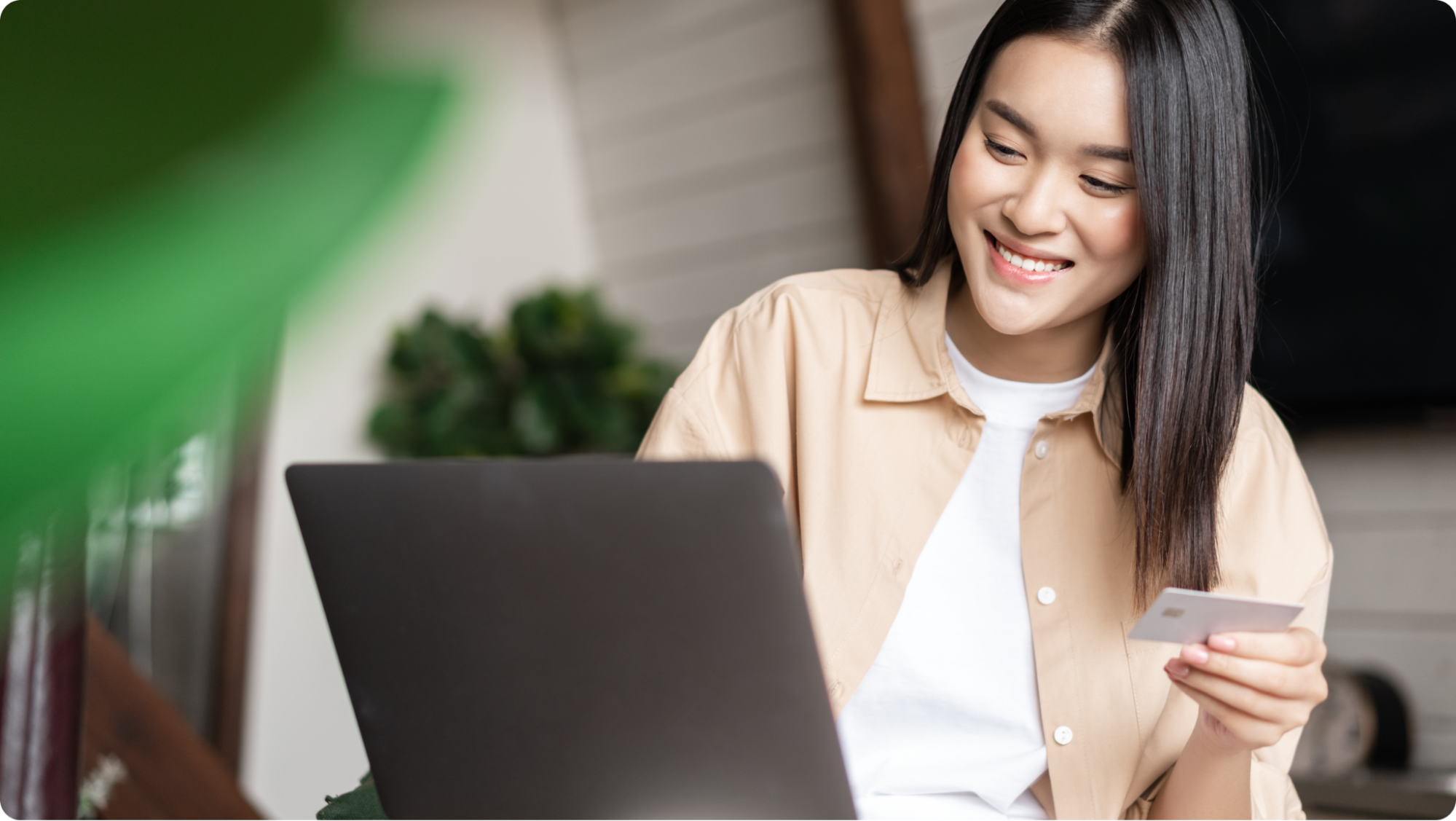 A woman smiling while looking at a laptop, holding a credit card in her hand, suggesting she is shopping or banking online in a cozy indoor setting.