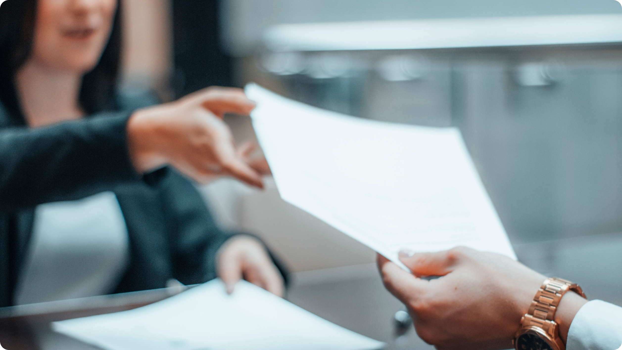 Two people sitting across a desk, one person handing a document to the other. The scene is out of focus, but both individuals appear to be in a professional setting.