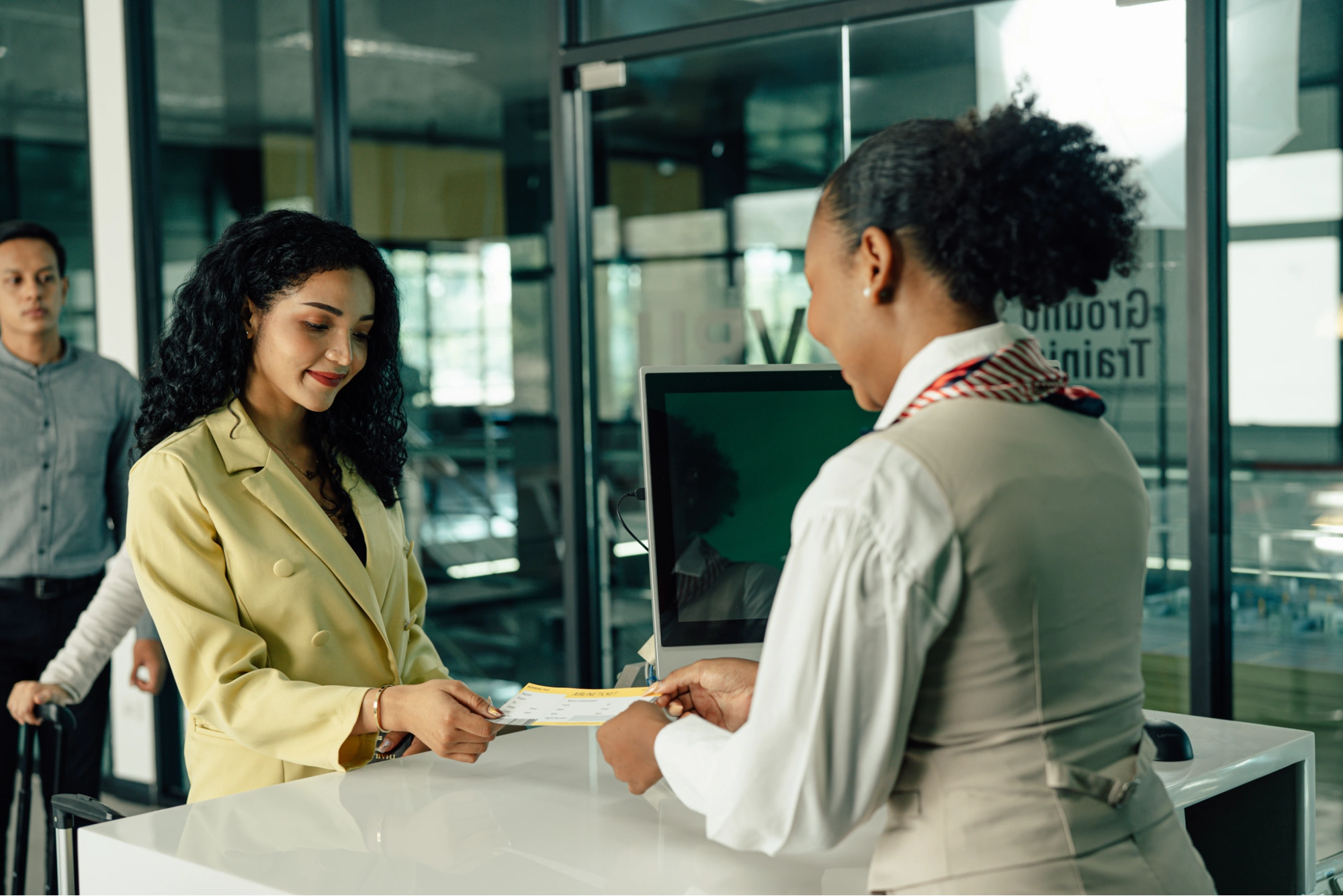 A woman in a yellow blazer smiles as she receives a boarding pass from an airline staff member at an airport check-in counter, perhaps contemplating business vs personal bank account options for her upcoming trip. Another passenger stands behind her with a suitcase.