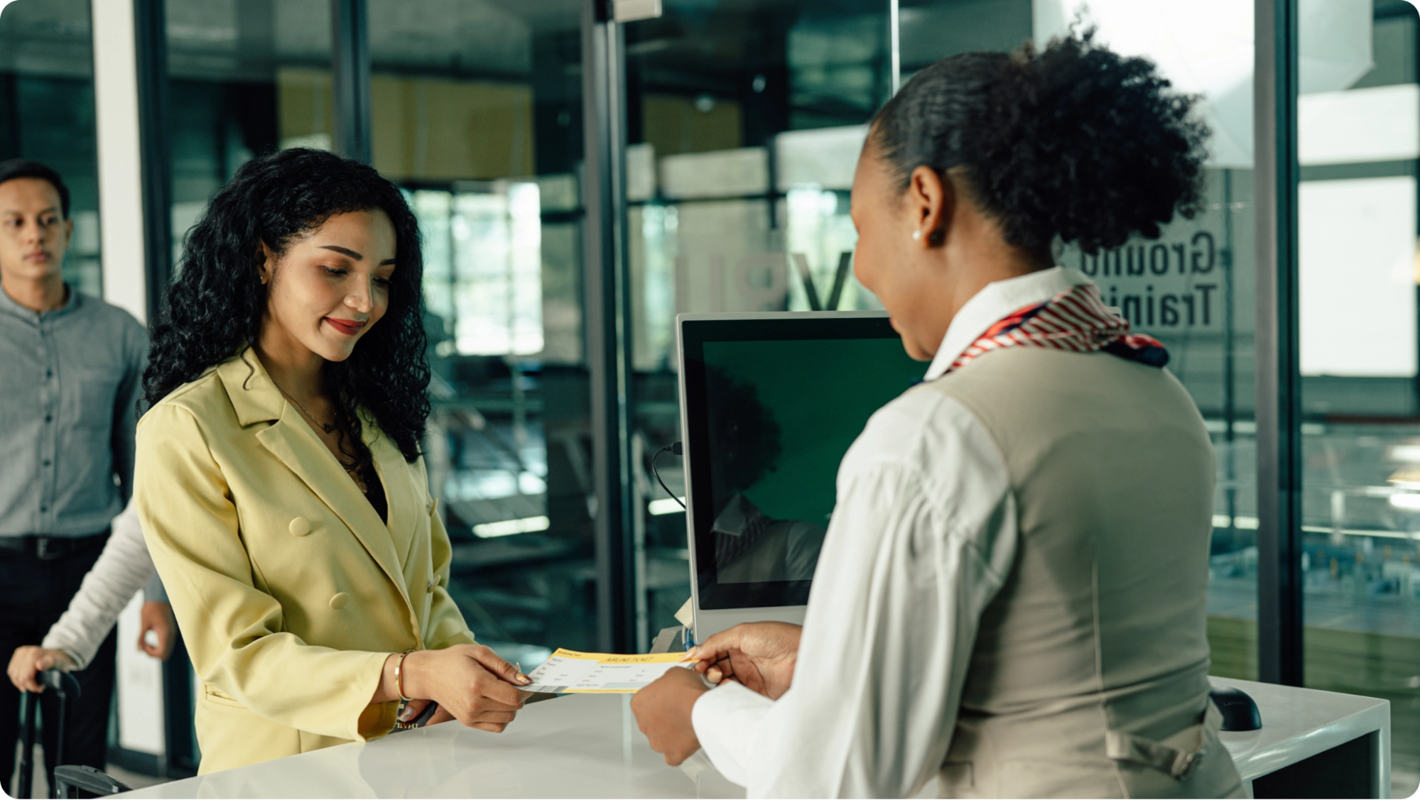 A woman in a yellow blazer smiles while handing a document to a receptionist at a modern office, possibly opening a business vs personal bank account. Another person stands in the background near sleek glass walls.