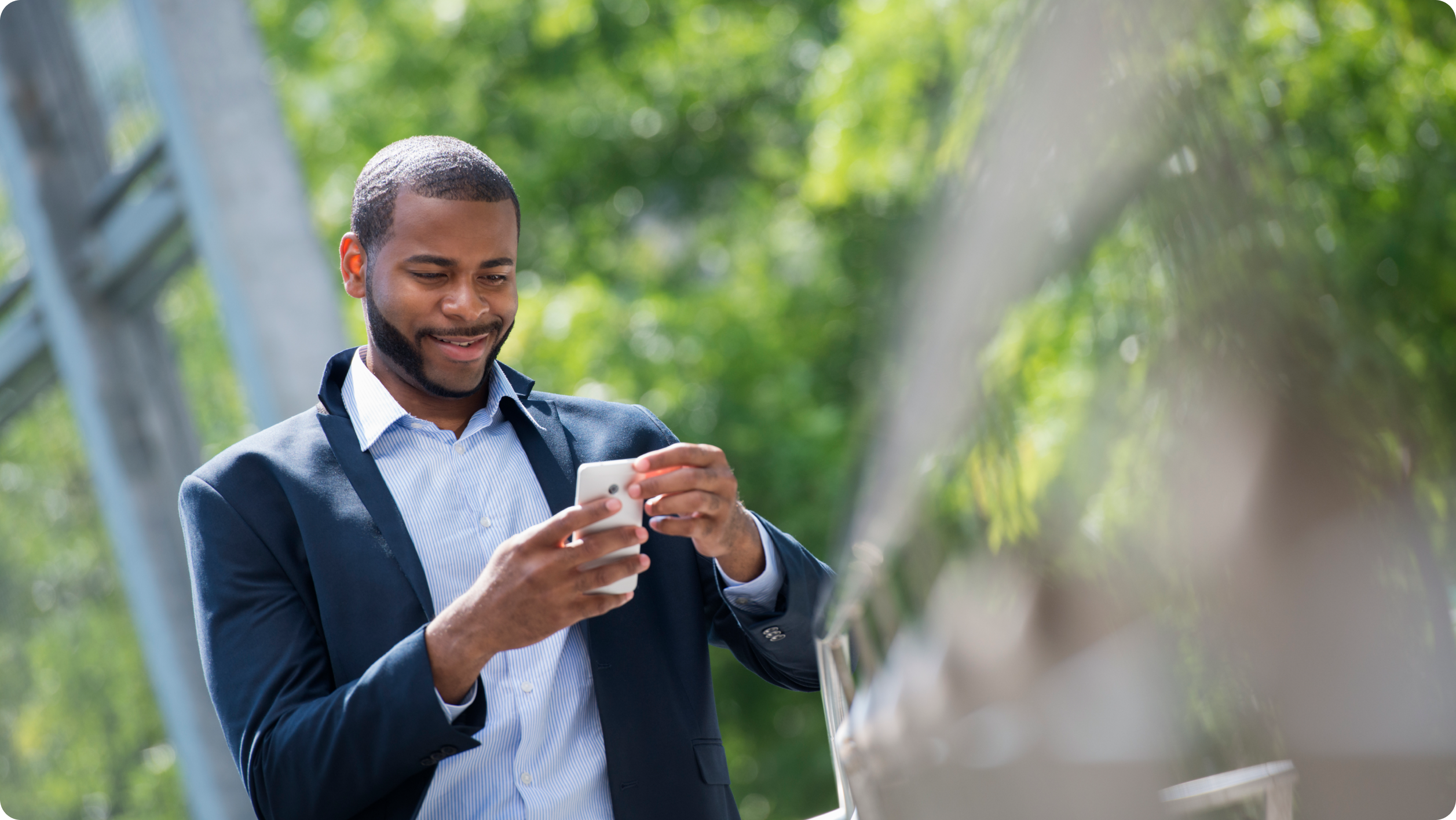 A man wearing a suit jacket and collared shirt stands outdoors, smiling as he looks at his smartphone—perhaps checking the latest top 10 rental property management software. Green trees and a metal structure are visible in the blurred background.