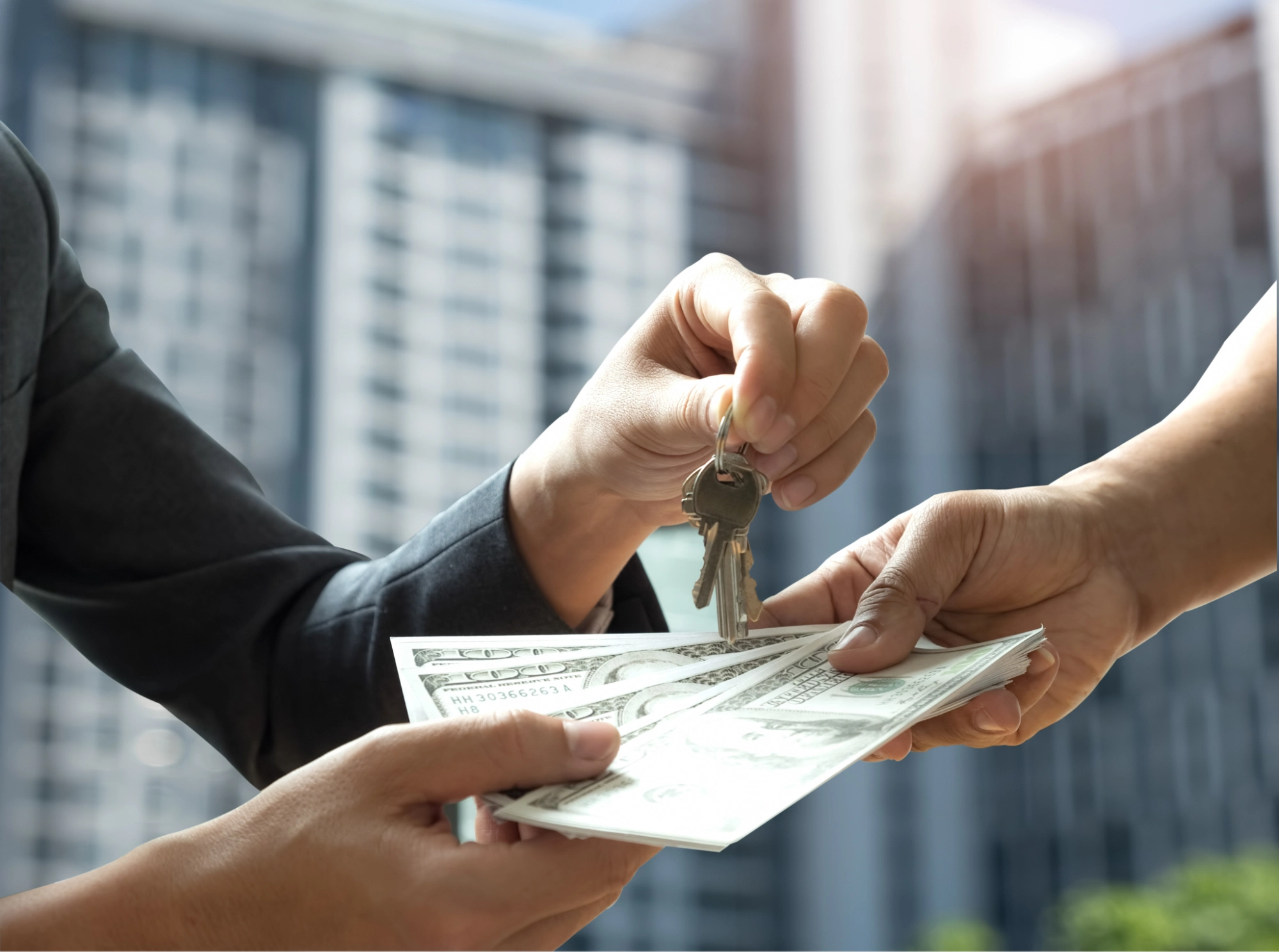 A person hands over a set of keys to another, who is offering a stack of hundred-dollar bills—perhaps sealing a deal that highlights the importance of keeping a business vs personal bank account​, with tall buildings blurred in the background.