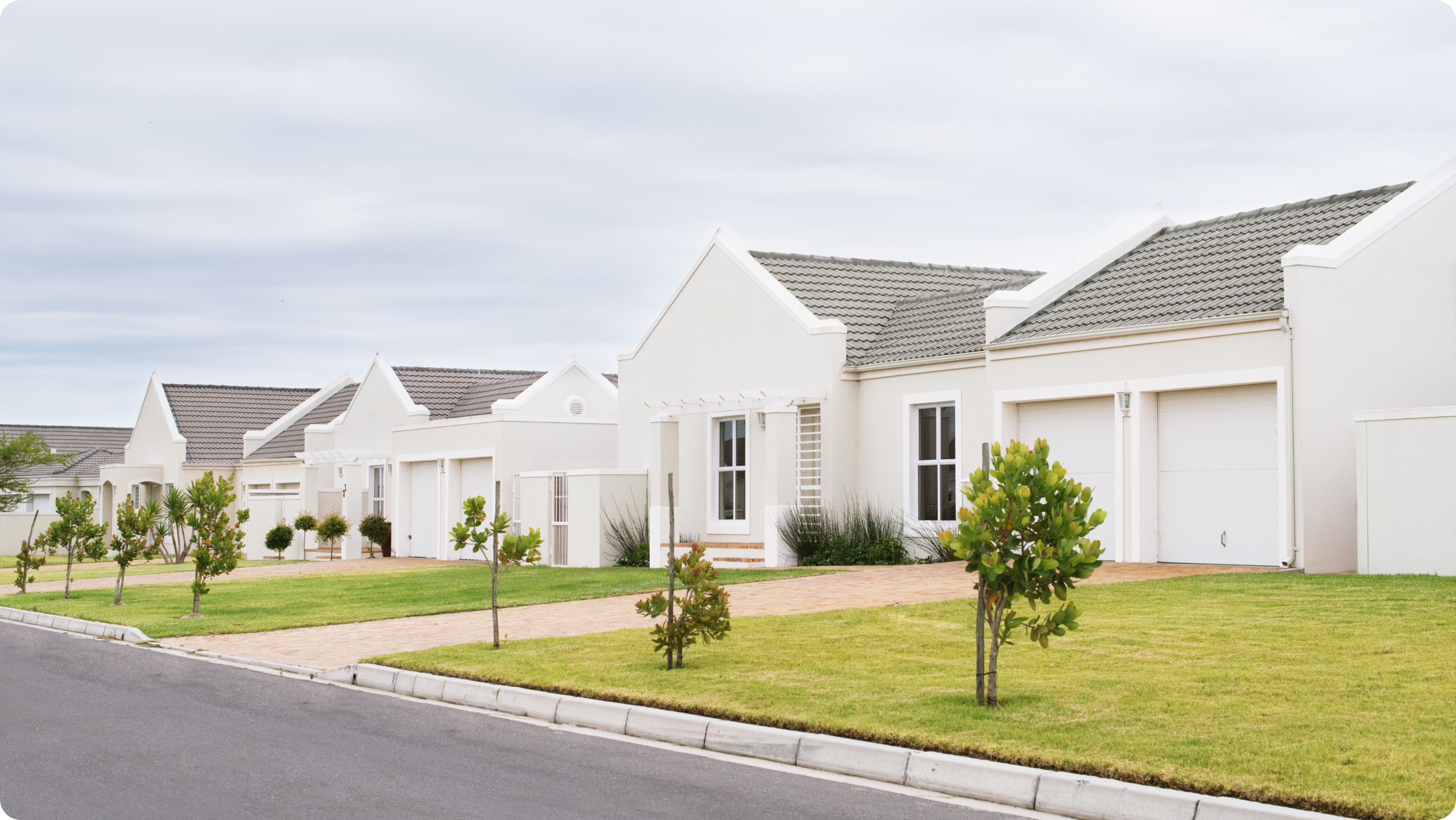 A row of modern, single-story white houses with gray roofs, neatly trimmed lawns, and small trees lines a quiet suburban street under a cloudy sky—perfect for pondering the differences between a business vs personal bank account​.