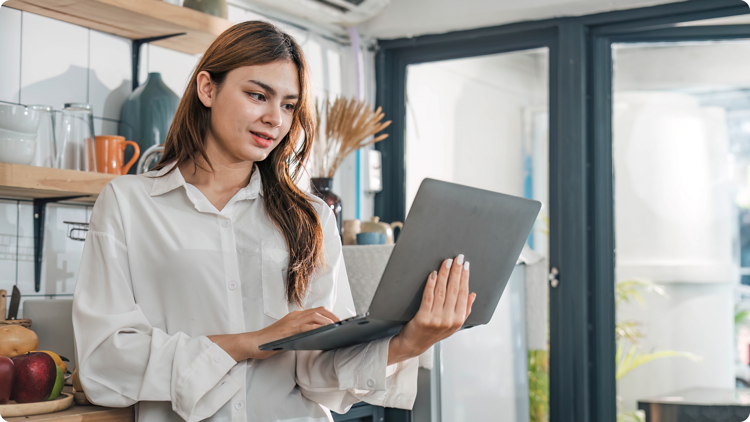 A woman in a white shirt stands in a modern kitchen, using her laptop to explore short term rental management software. She smiles at the screen, surrounded by kitchenware, shelves, and glass doors letting in natural light.