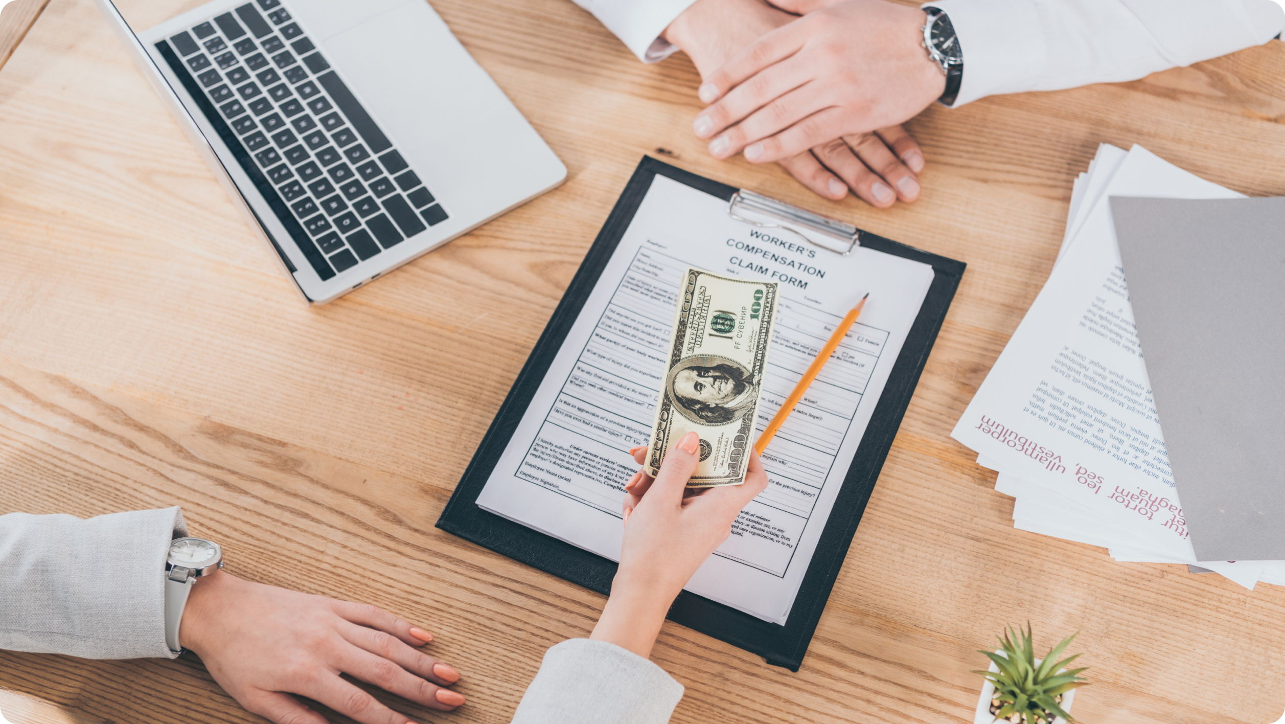 A person hands a stack of hundred-dollar bills to another across a desk with a laptop displaying rental management software, surrounded by documents and a clipboard holding a "Worker’s Compensation Claim Form.