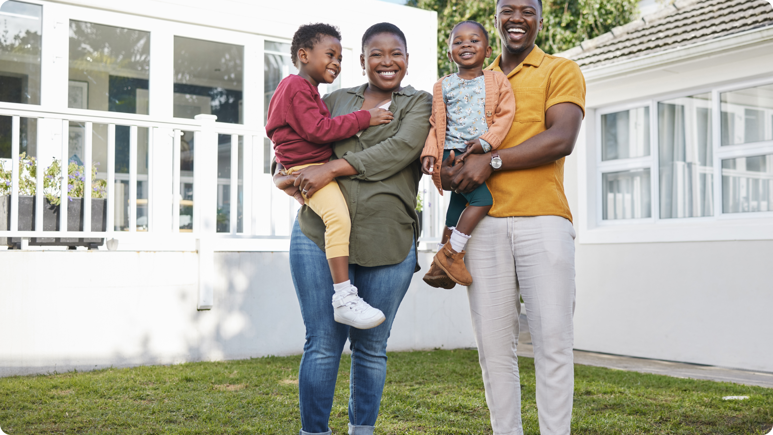 A smiling family of four stands outside their house, enjoying time together in the front yard. The parents each hold a young child in their arms, all dressed in colorful clothing—peaceful and content thanks to trusted HOA bank services.