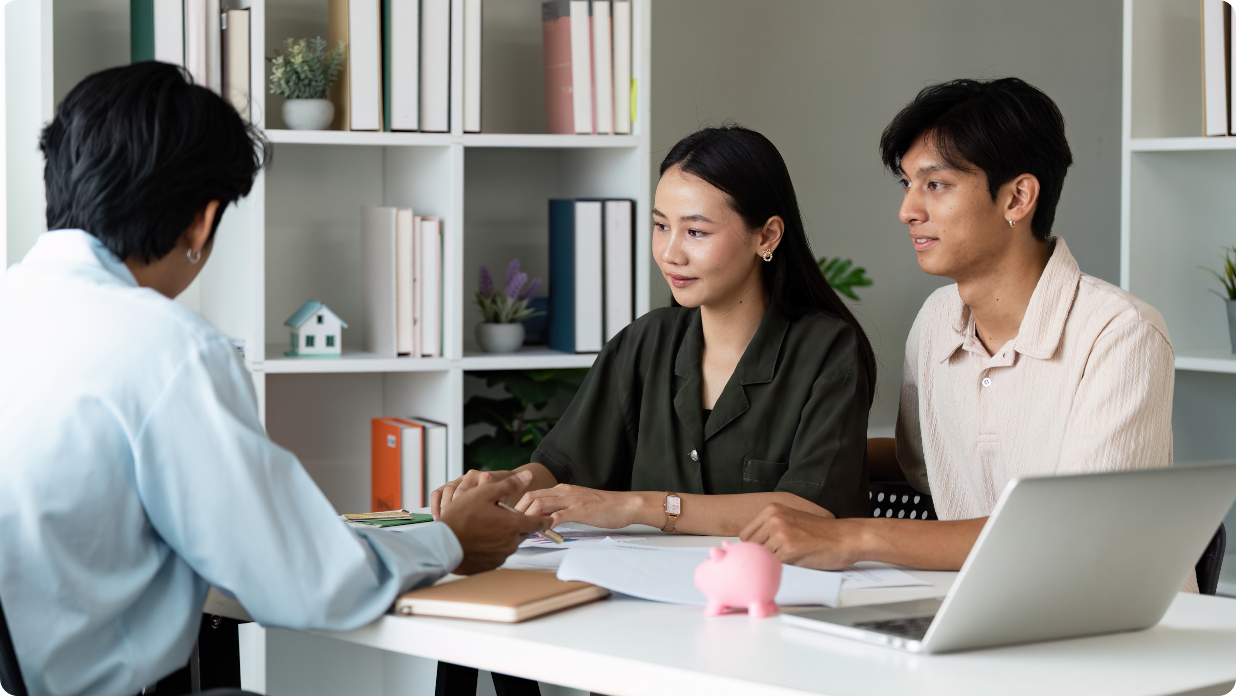 A couple sits at a desk discussing bank options and loans with a professional. Papers, a notebook, a pink piggy bank, and a laptop are on the desk. Bookshelves with files and decor are visible in the background.