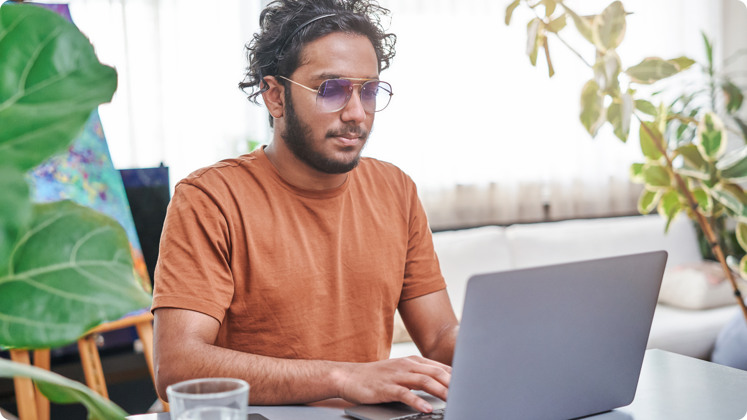A man wearing sunglasses and a brown t-shirt works on his laptop at a table, possibly researching business vs personal bank account options, in a bright room with plants and a glass of water nearby.