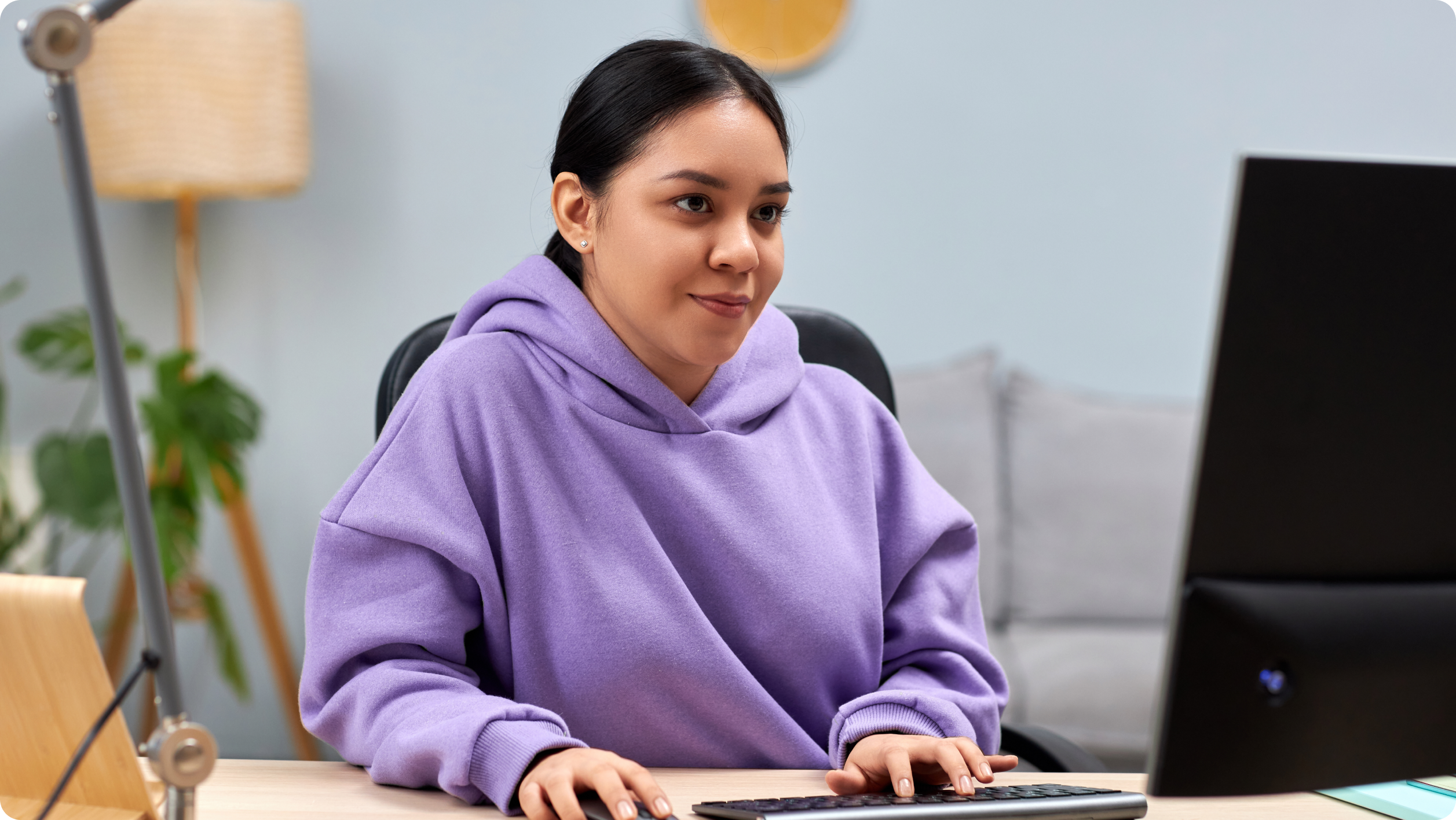 A woman in a purple hoodie sits at a desk, focused on her computer as she researches how to open an escrow account. She is indoors, with a lamp and a couch in the background.