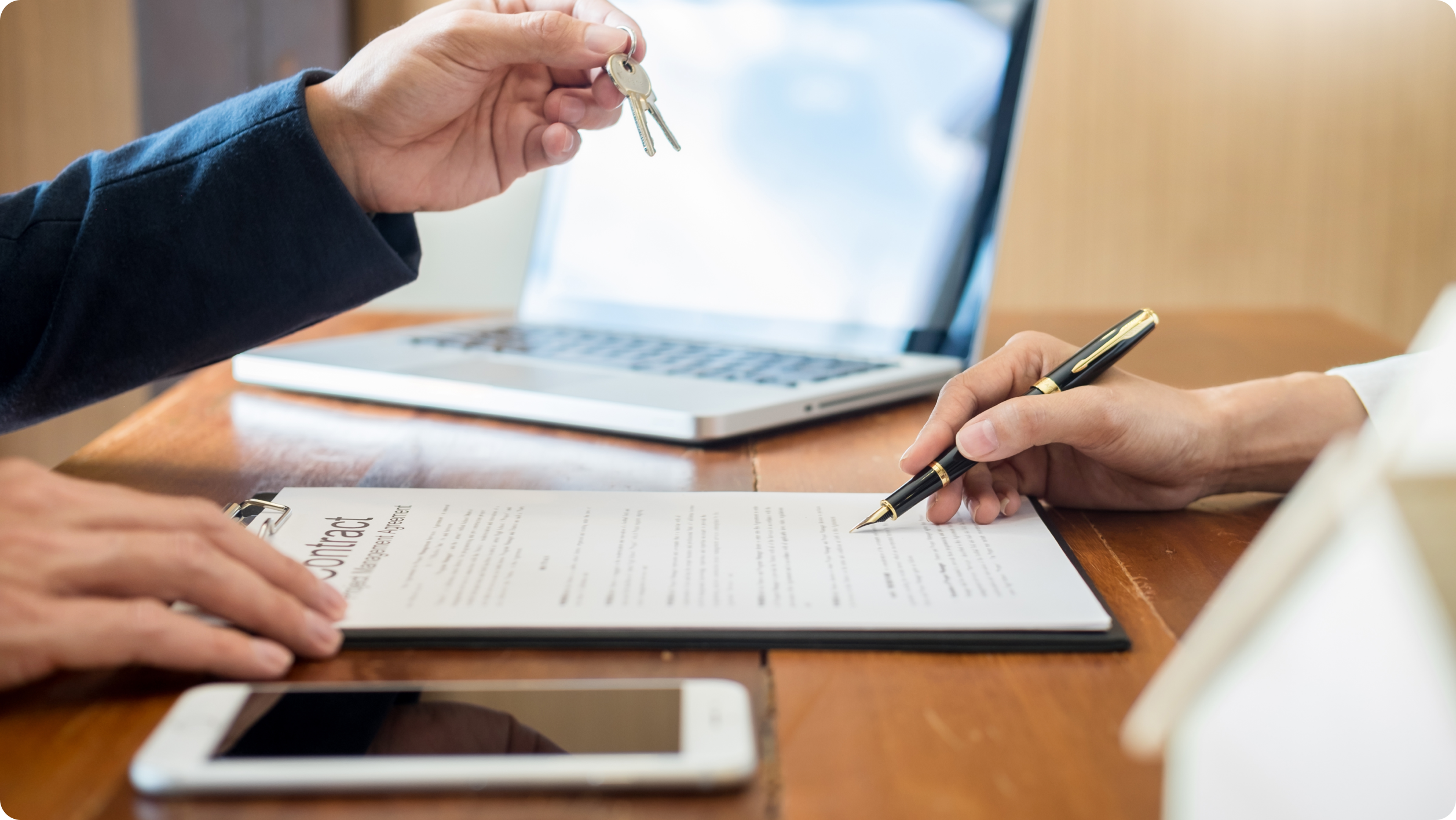 A person hands over house keys to another person who is signing a contract at a table, with a laptop and smartphone nearby—highlighting the importance of distinguishing between a business vs personal bank account​ in property transactions.