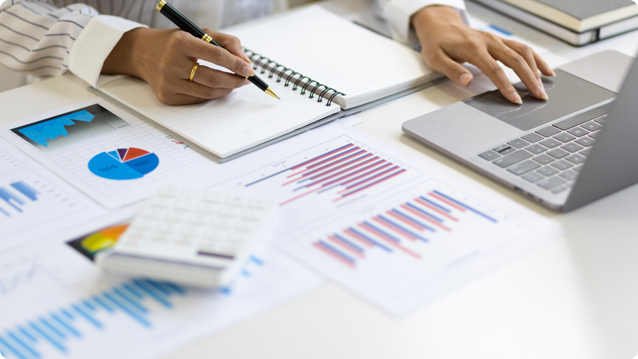 A person writes in a notebook while using a laptop, surrounded by charts, graphs, and a calculator on a desk—suggesting financial analysis or business planning involving HOA bank services.