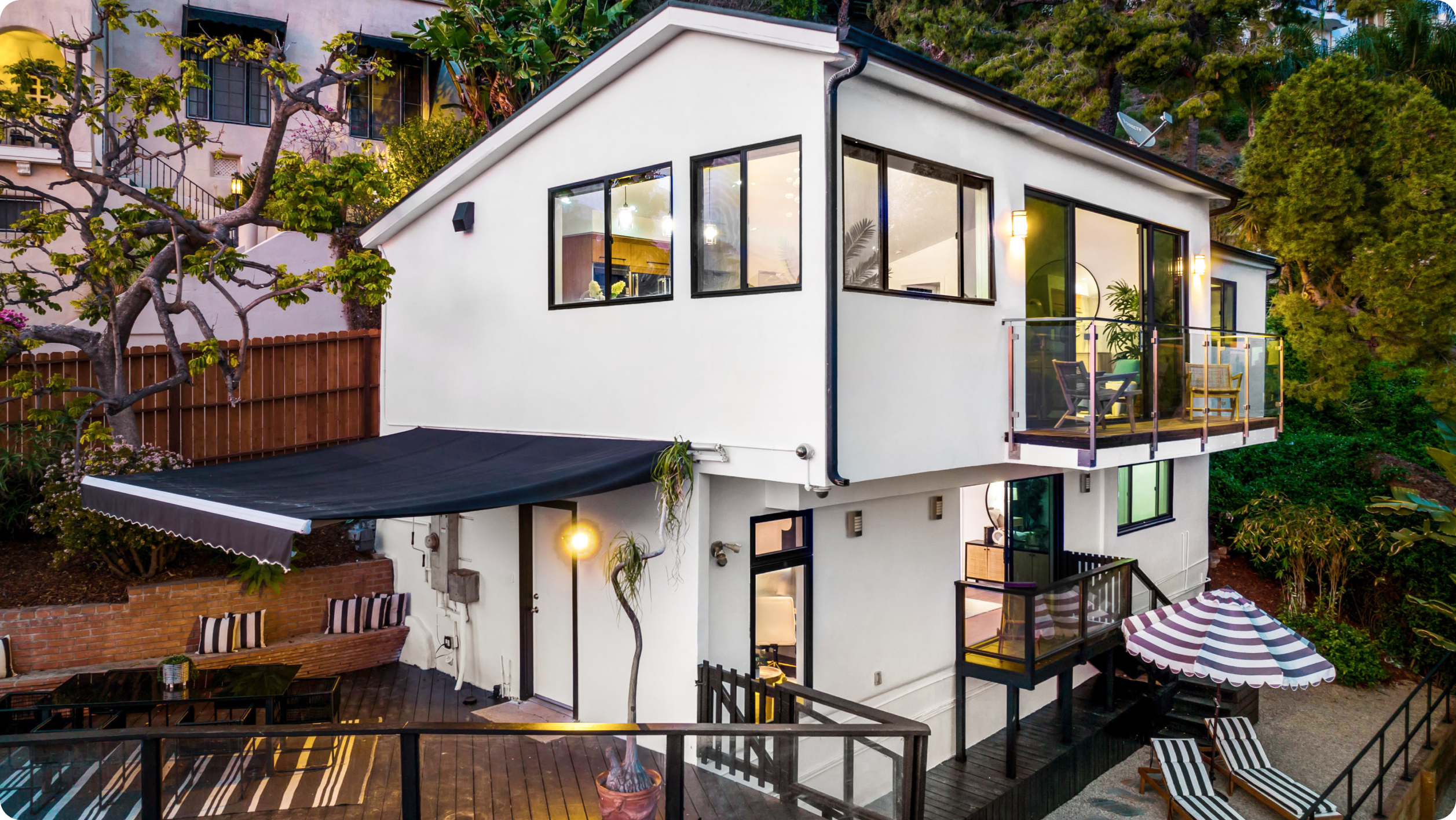 Modern two-story white house with large windows, a balcony with seating, outdoor patio areas, black railings, and a retractable awning—an ideal retreat for those who value real estate financial management—surrounded by greenery and trees on a hillside.