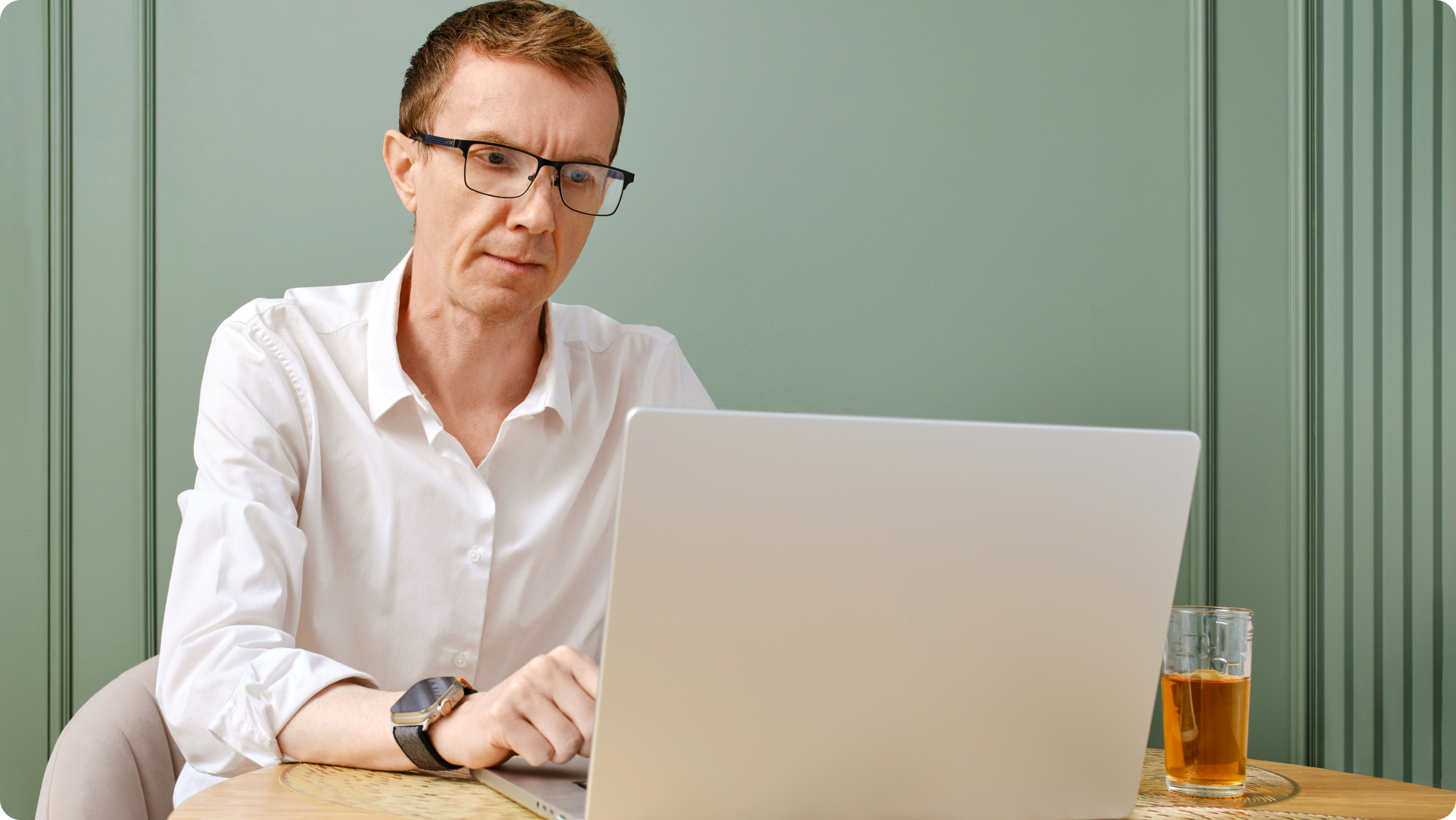 A man wearing glasses and a white shirt sits at a table working on his laptop, possibly exploring rent management software. A glass of tea rests beside him, with a green wall and decorative molding in the background.