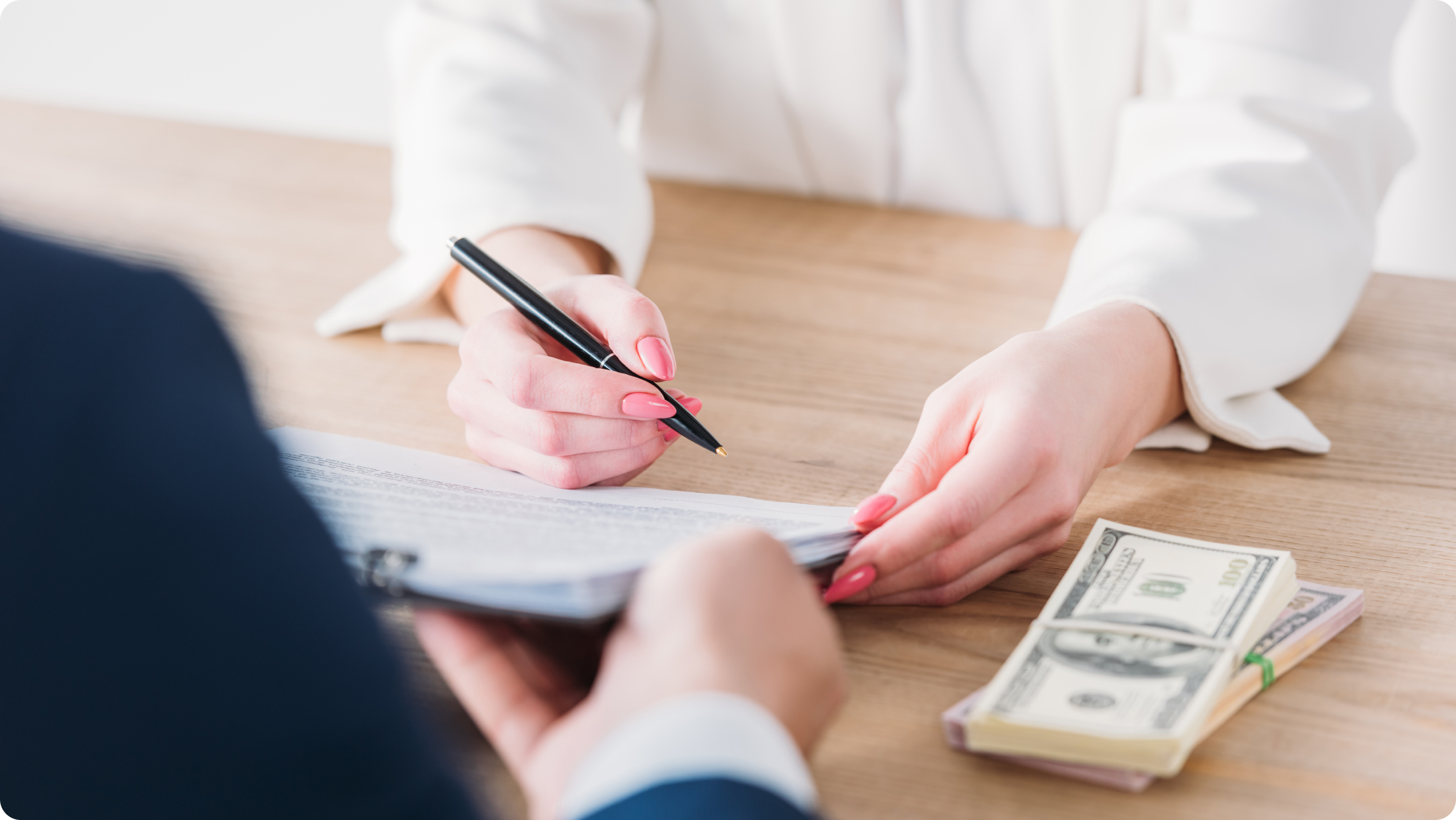 A woman with pink nails is signing a document on a desk, with a stack of U.S. dollar bills beside her—suggesting an agreement involving hoa bank services. Another person is holding the document for her.