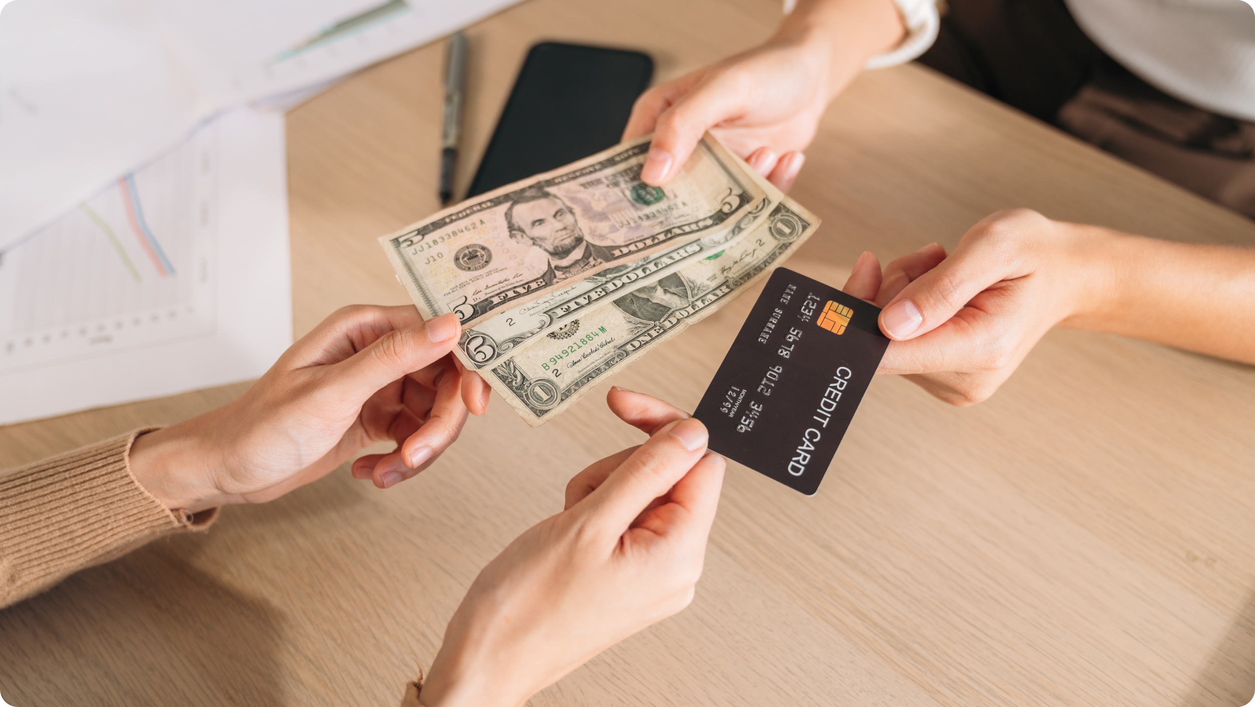 Two people exchange payment, with one hand holding cash and the other a credit card—illustrating choices faced when managing business vs personal bank account​ transactions over a wooden table scattered with documents and a smartphone.