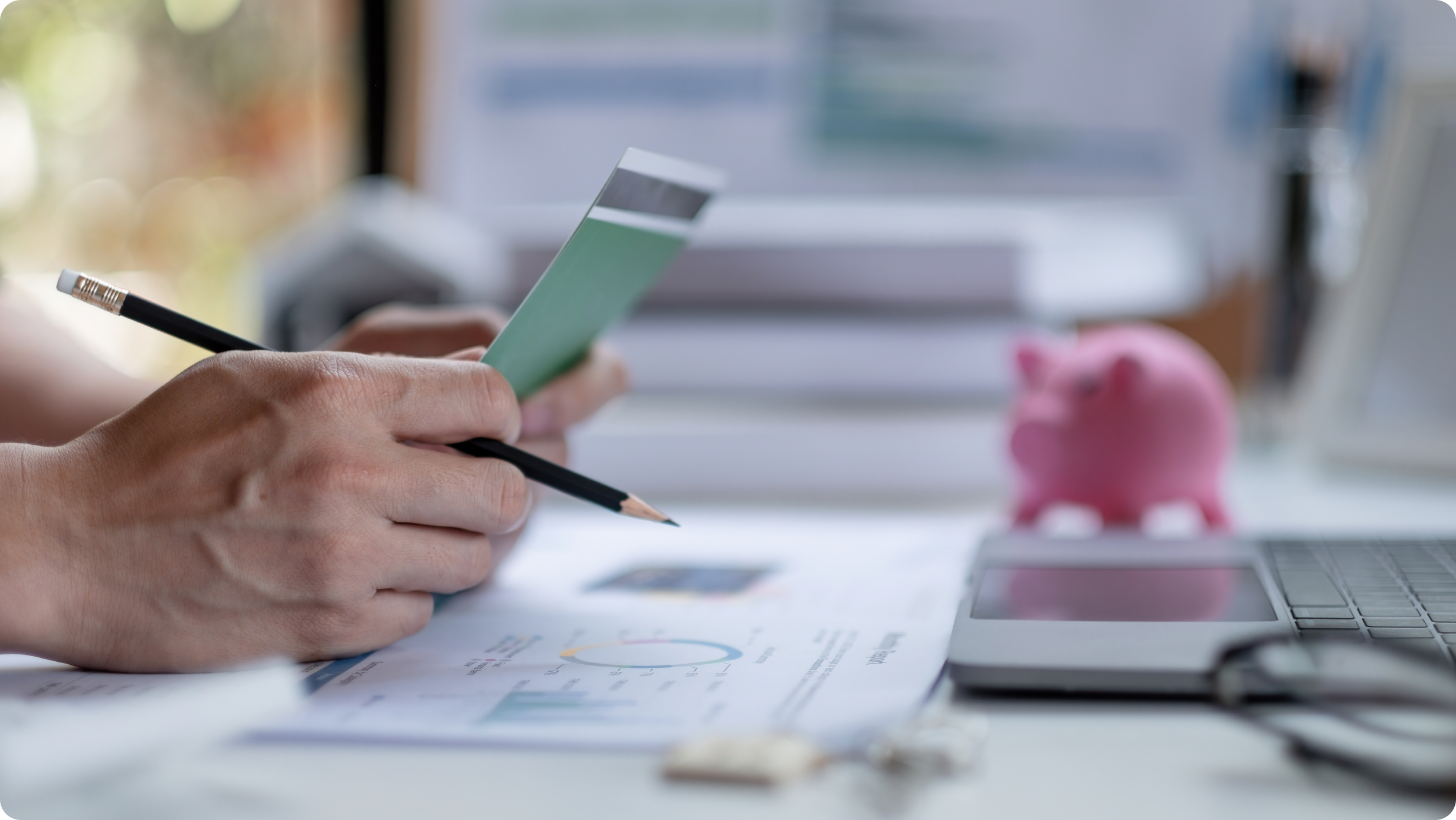 Close-up of a person holding a checkbook and pencil, working with financial documents at a desk. A pink piggy bank, laptop, and paperwork highlight budgeting while weighing business vs personal bank account​ options.