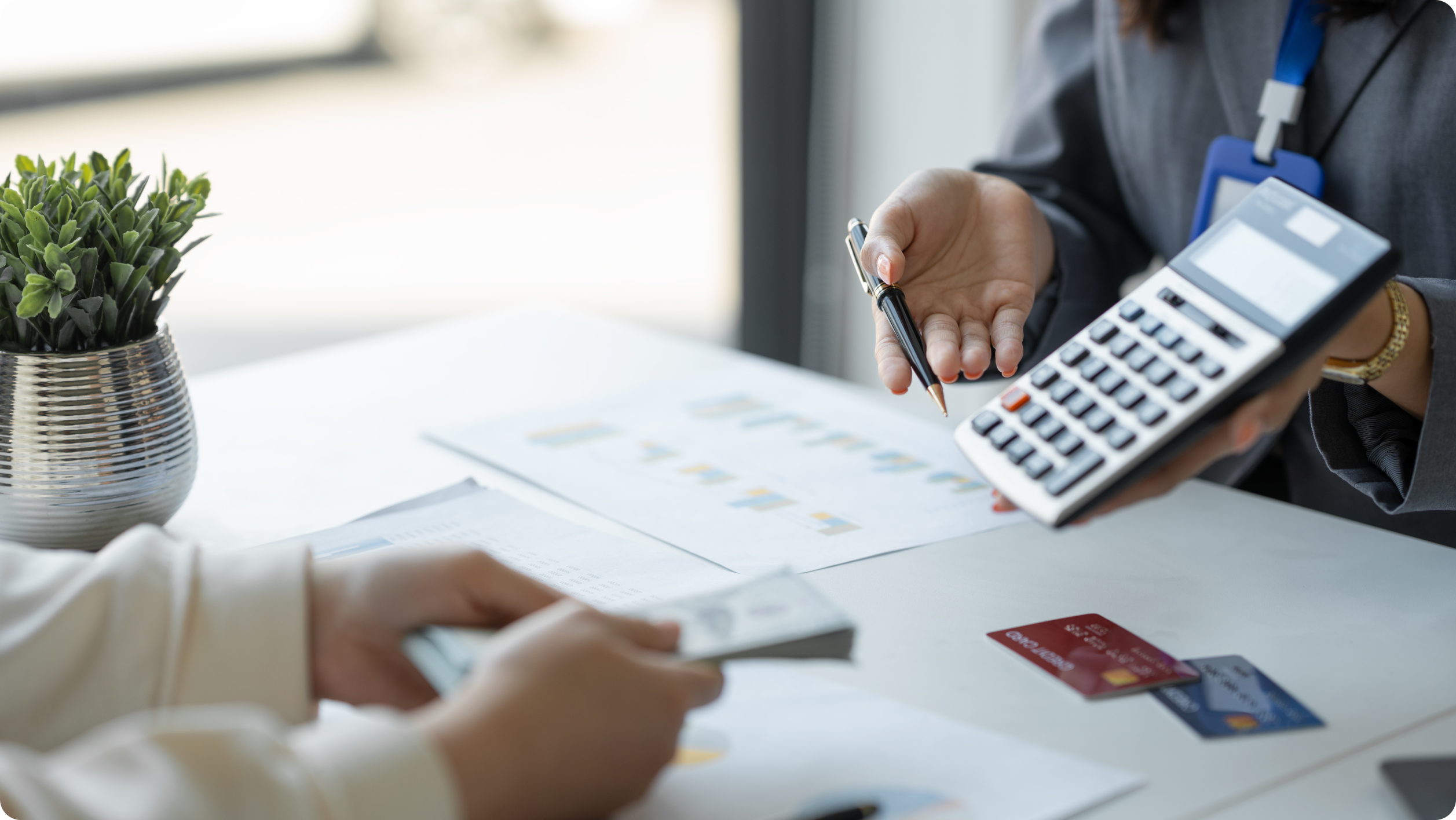 Two people sit at a desk with financial documents, a calculator, and credit cards, discussing business vs personal bank account matters. One holds cash while the other gestures with a pen, highlighting the importance of organized payments. A small potted plant decorates the desk.