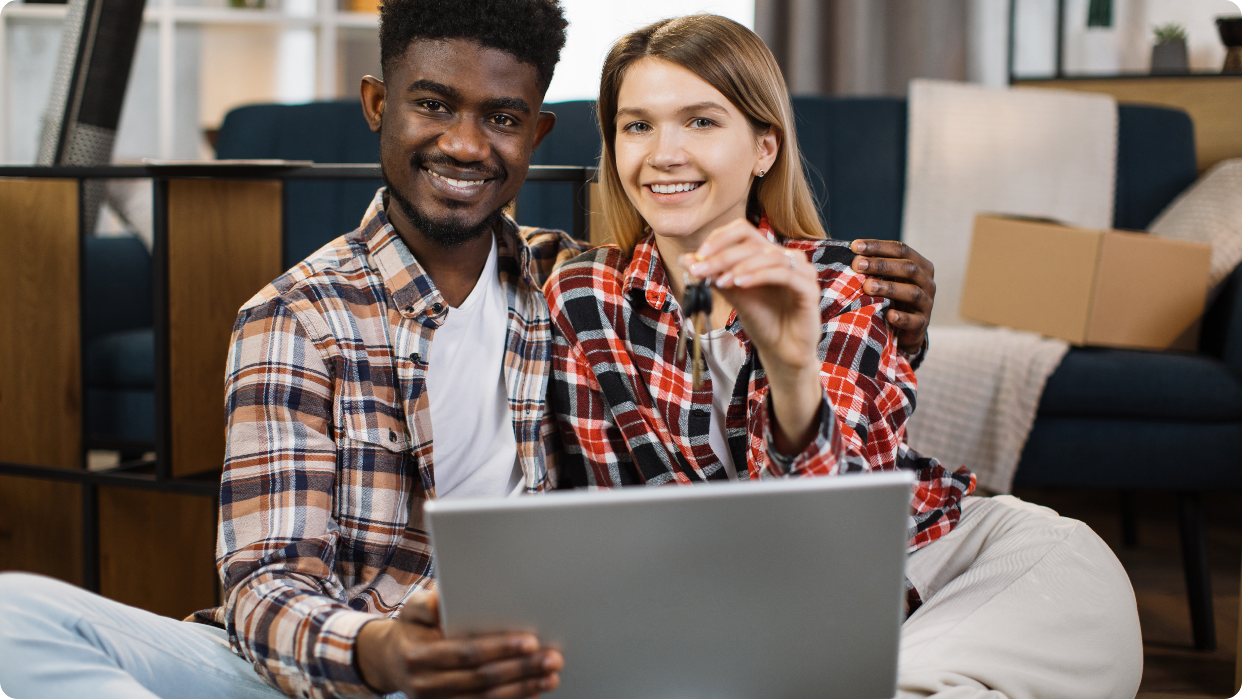 A smiling couple sits on the floor at home, looking at a laptop together. They appear happy and relaxed, perhaps exploring rent management software, with cardboard boxes and cozy furnishings visible in the background.