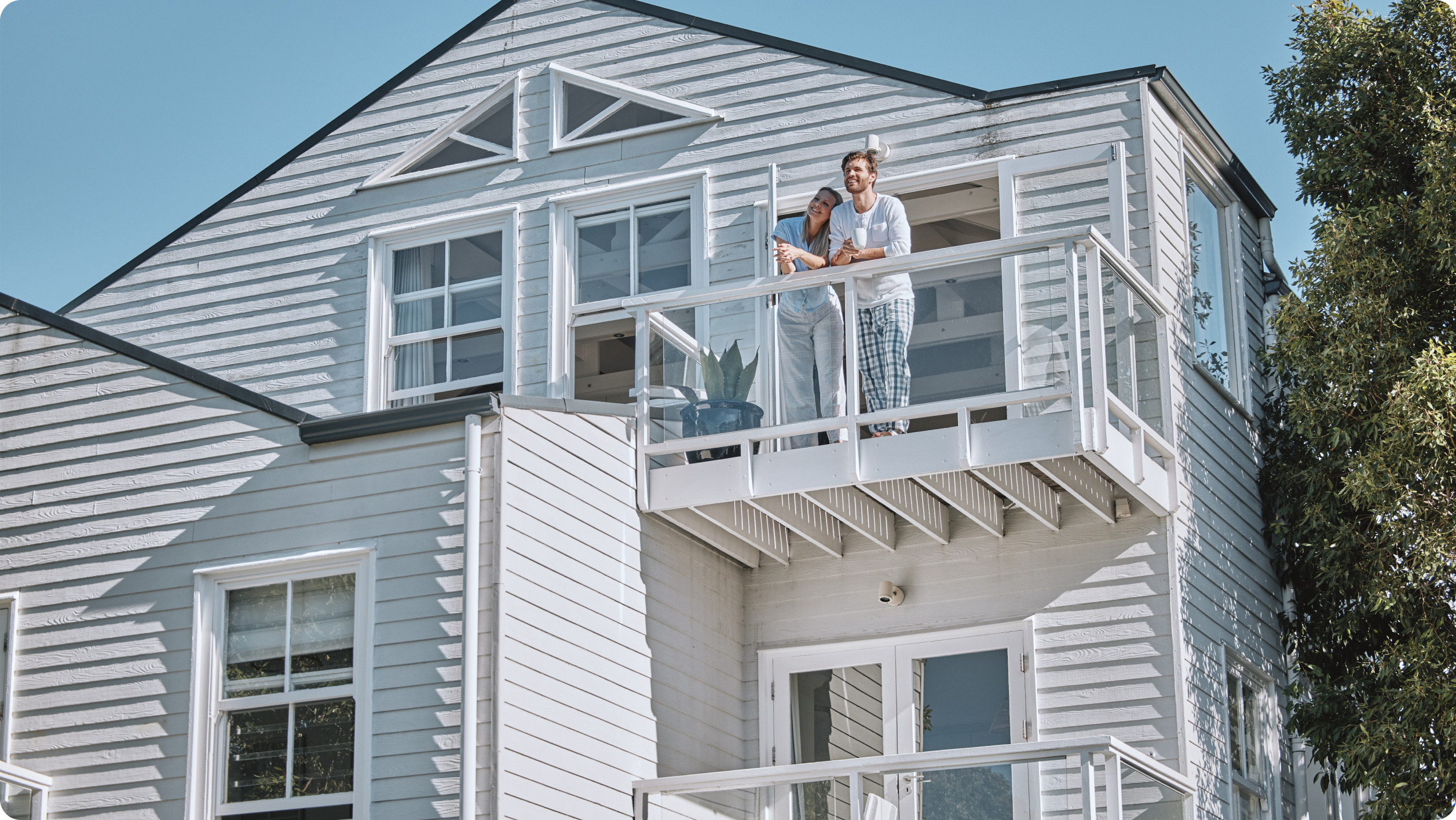 A couple stands together on the balcony of a modern white house with large windows, enjoying the sunny day. The property management banking team ensures everything runs smoothly as they take in views of trees under a clear blue sky.