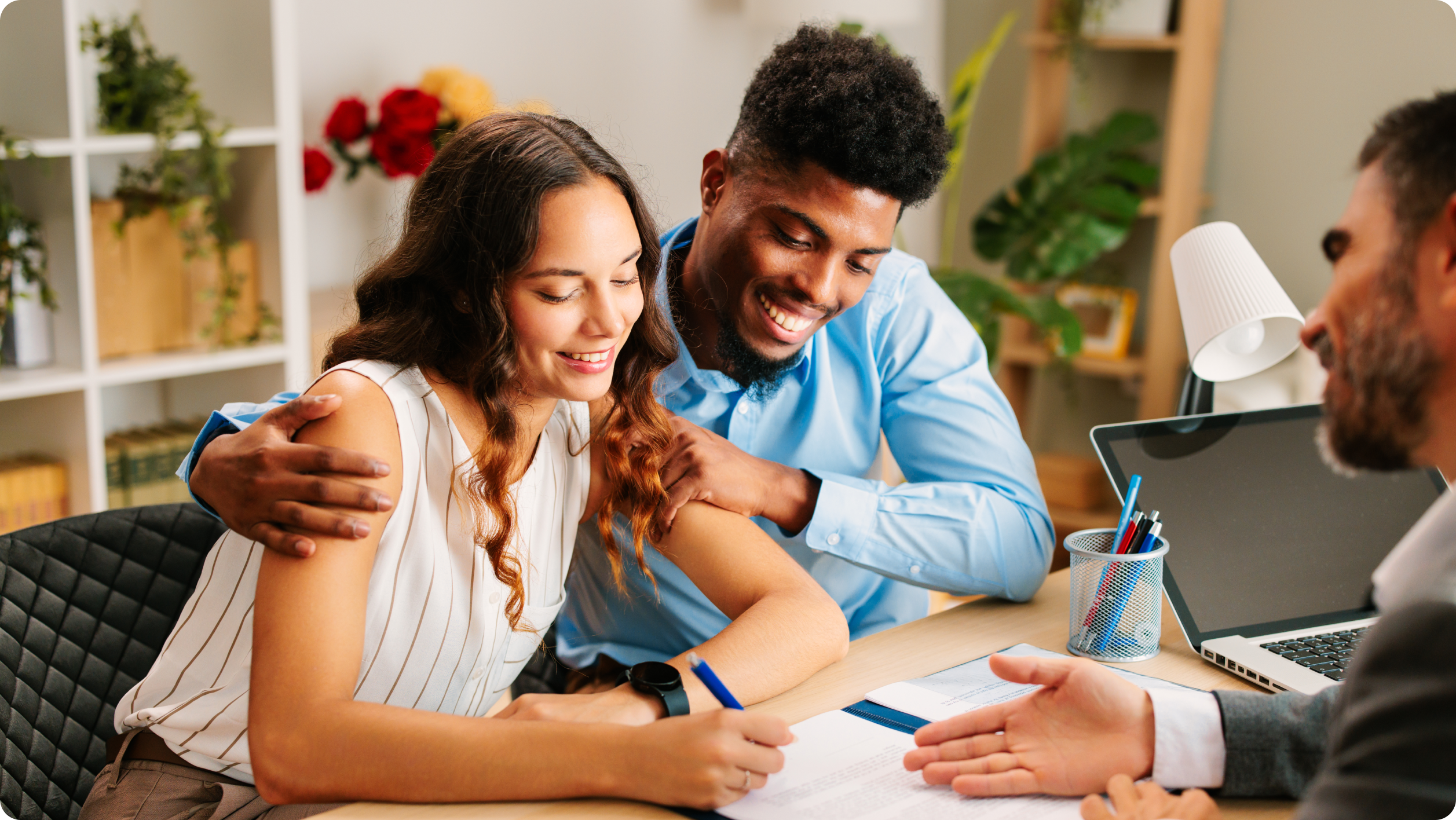 A smiling couple sits at a desk, the woman signing a document while the man embraces her. Across from them, someone gestures to the paper—highlighting hoa bank services​ in what appears to be a business or financial agreement.