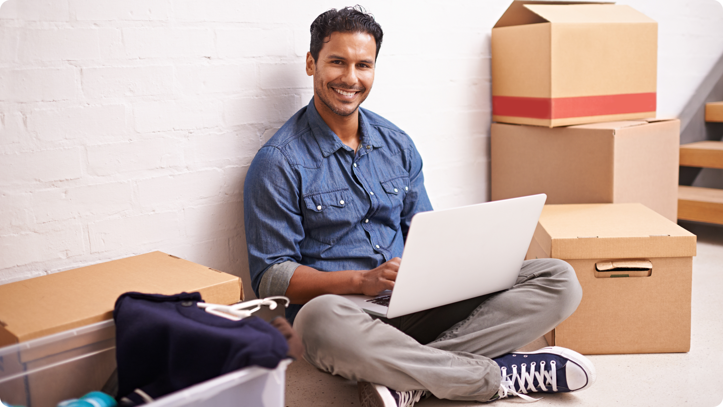 A man sits on the floor next to moving boxes and a plastic bin, smiling at the camera while using a laptop. He is dressed in a blue shirt and khaki pants, suggesting he is in the process of moving.