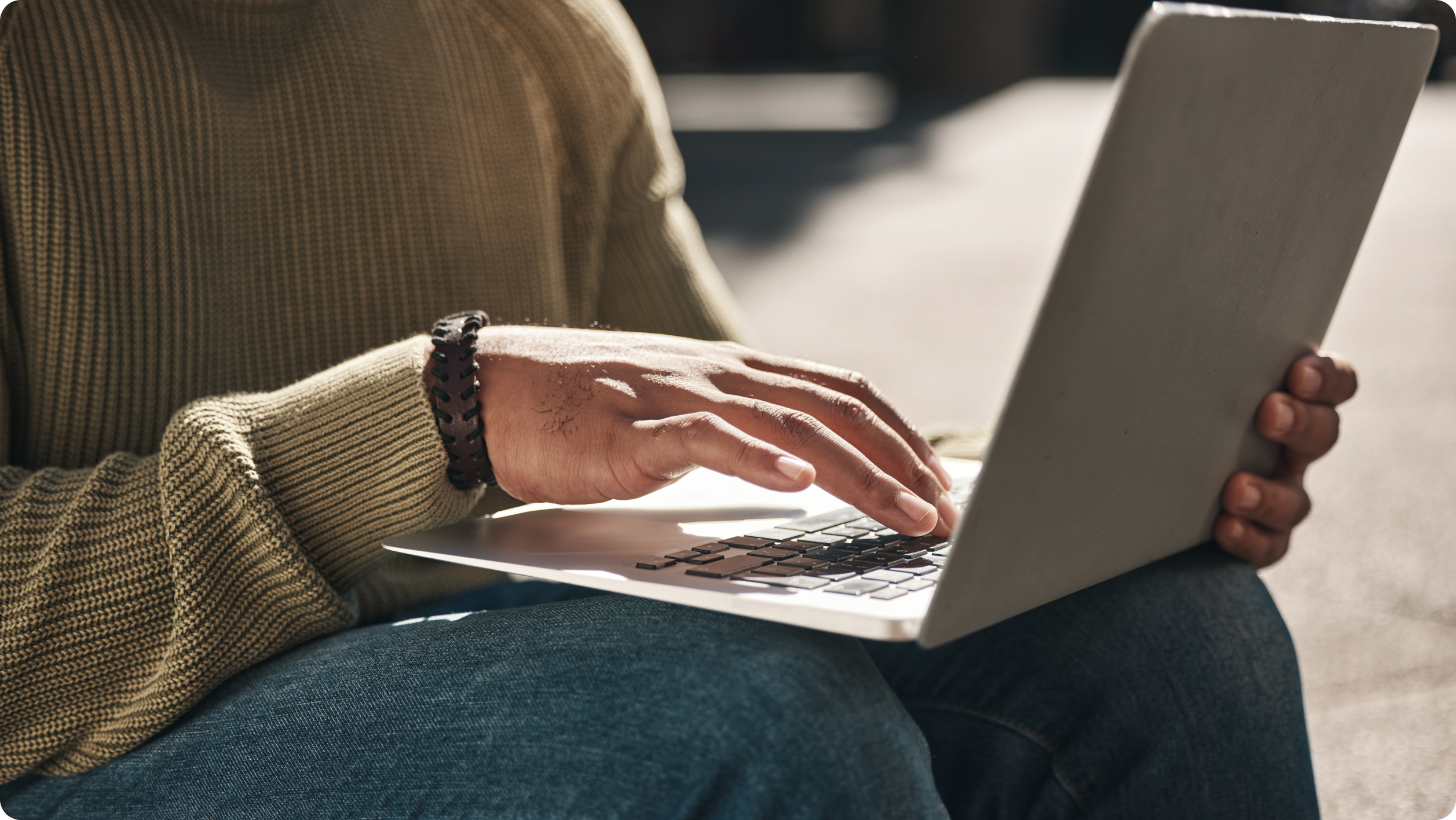 A person wearing a green sweater and blue jeans is sitting and typing on a laptop, possibly working on real estate financial management tasks. Only their hands and part of their body are visible.