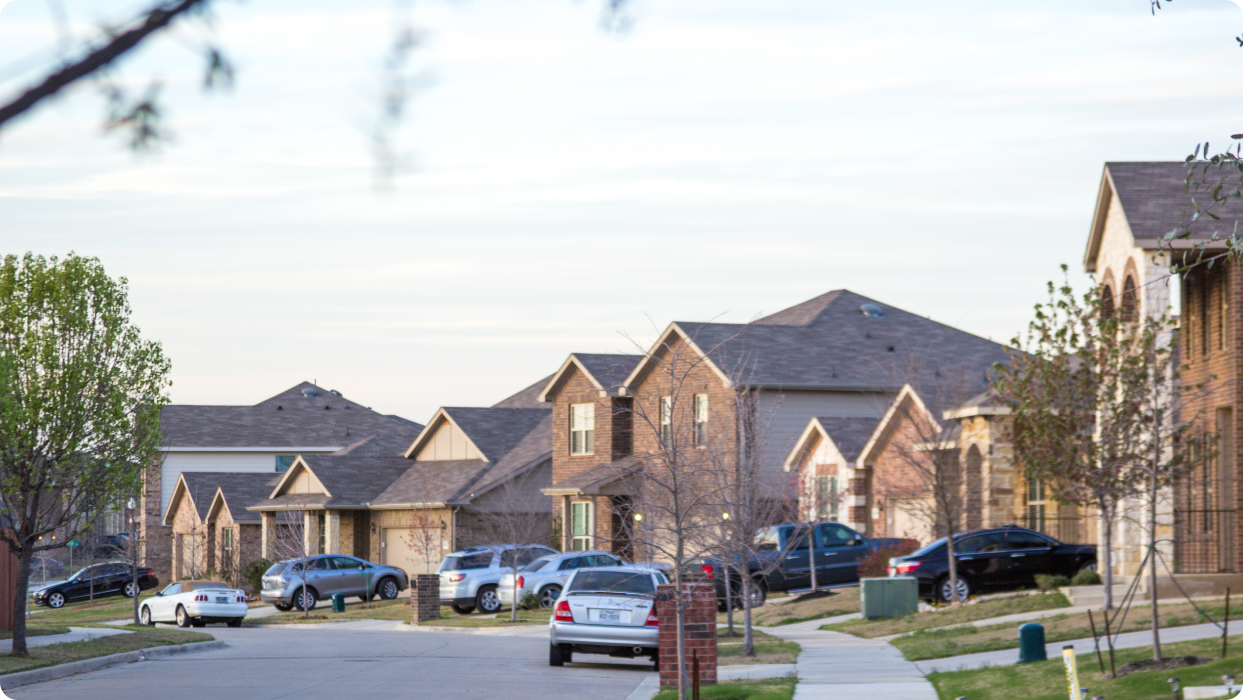 A suburban street lined with houses, cars parked by the curb, and trees with sparse green leaves on a clear day—much like considering business vs personal bank account​ options for managing your finances in a vibrant community.