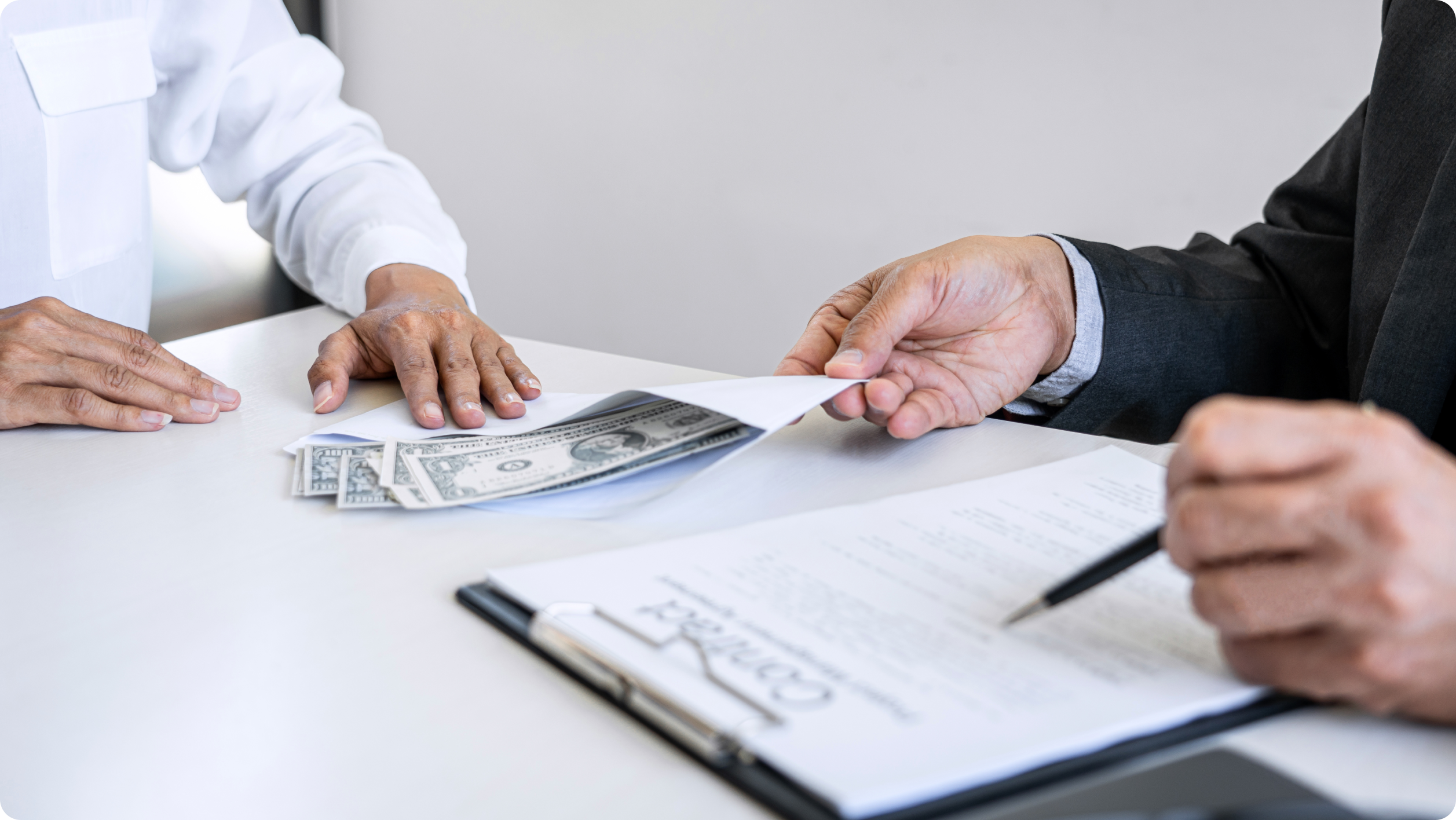 A person in a suit hands an envelope containing cash to another person across a table, with a contract, pen, and rental management software open on a nearby laptop.