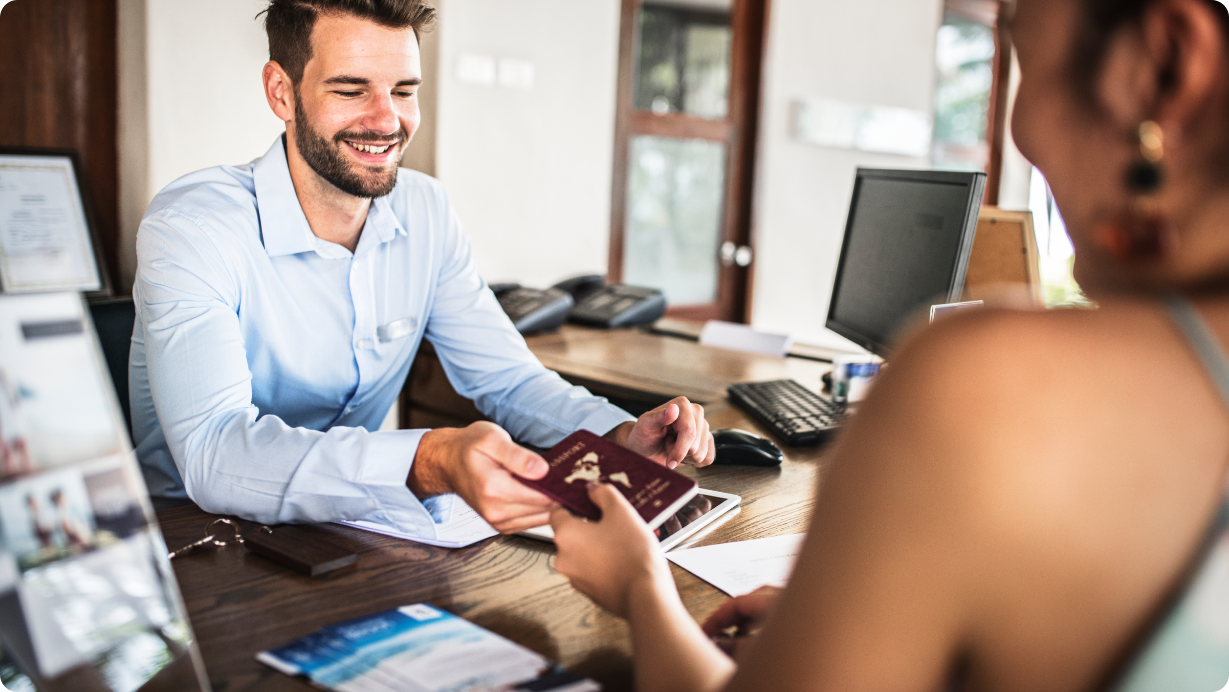 A smiling man in a blue shirt hands a passport to a woman across a desk in a bright office, possibly at a travel agency or hotel reception, where brochures and computers sit alongside leaflets about business vs personal bank accounts.