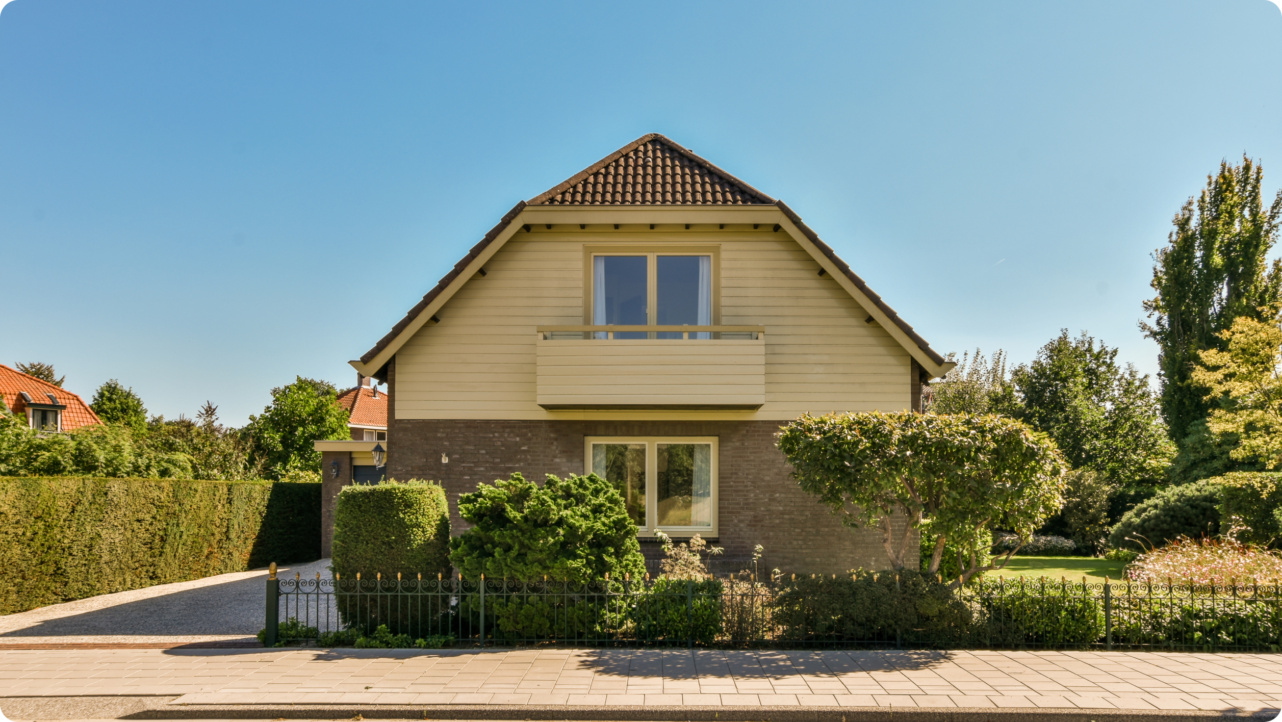 A two-story house with a sloped red-tiled roof, light-colored upper walls, brick lower walls, large windows, and a small balcony surrounded by greenery—perfect for enjoying the sun and convenient access to HOA bank services​.