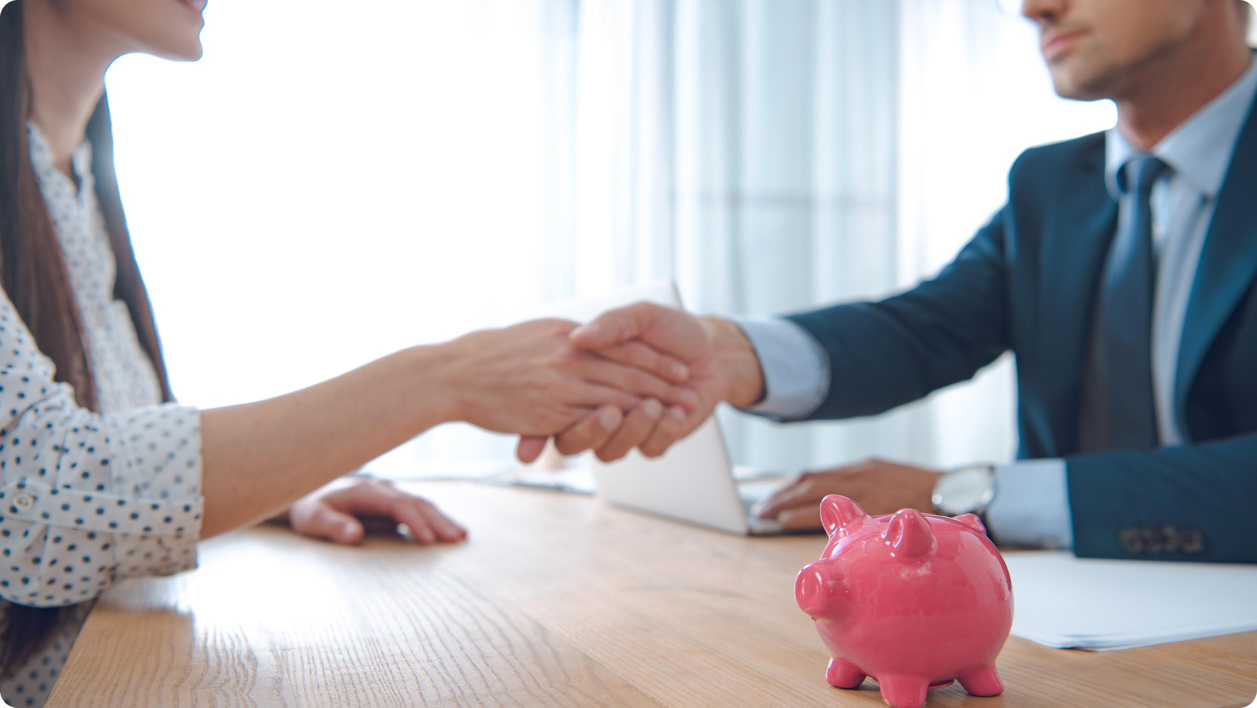 Two people in business attire shake hands across a desk with a pink piggy bank and a laptop, suggesting an agreement on bank options or loans.