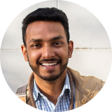 A young man with short dark hair and a beard smiles at the camera. He is wearing a checkered shirt and a brown jacket, standing in front of a light-colored wall.