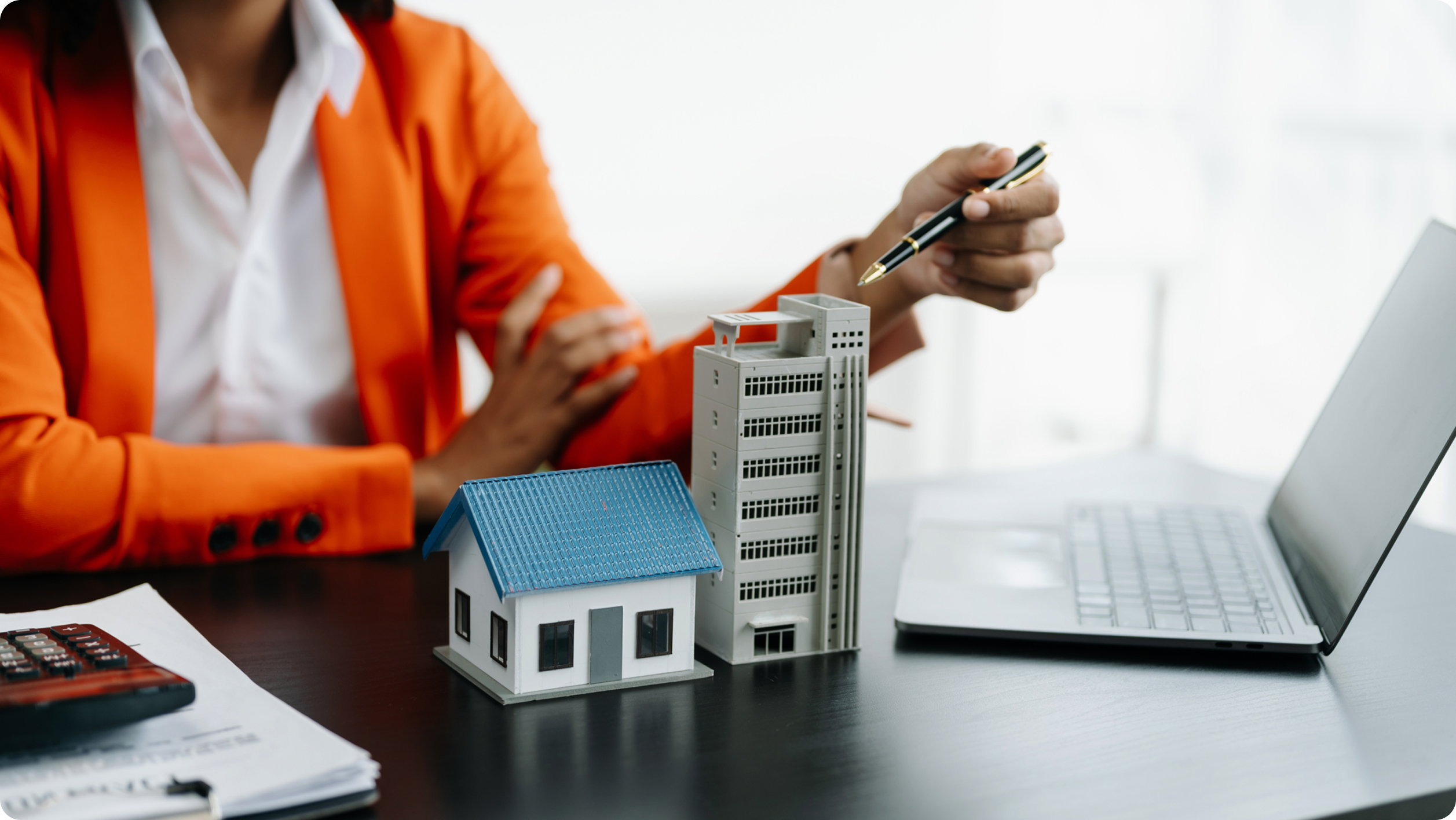 A person in an orange blazer gestures with a pen near miniature models of a house and an apartment building on a desk, next to a laptop and documents, perhaps discussing how to get a high yield savings account for future investments.