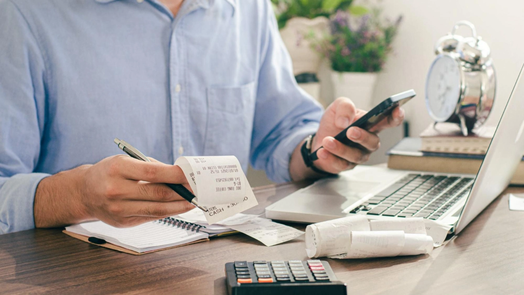A person sits at a desk with a laptop, holding receipts and a pen in one hand and a smartphone in the other. There is a notebook, calculator, alarm clock, and more receipts on the desk.