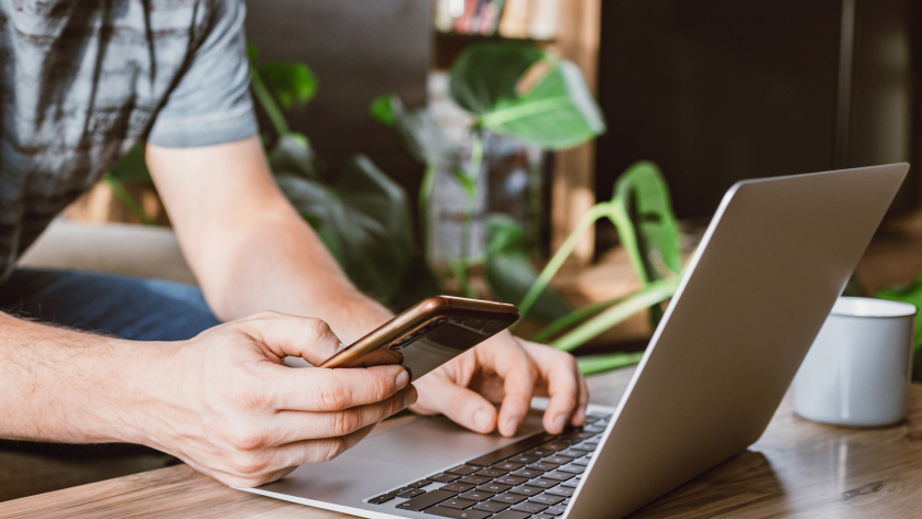 A person manages property management banking tasks, using a smartphone with one hand and typing on a laptop with the other, seated at a wooden table indoors with a green plant and a mug in the background.