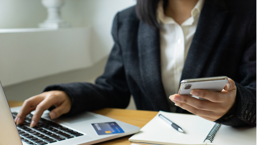 A person in a business suit uses a laptop with a credit card on the keyboard and holds a smartphone in one hand, with an open notebook and pen on the desk.