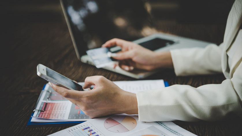 A person in a suit works at a desk with a laptop, holding a credit card and using a smartphone. Papers with charts and graphs are spread out on the desk.