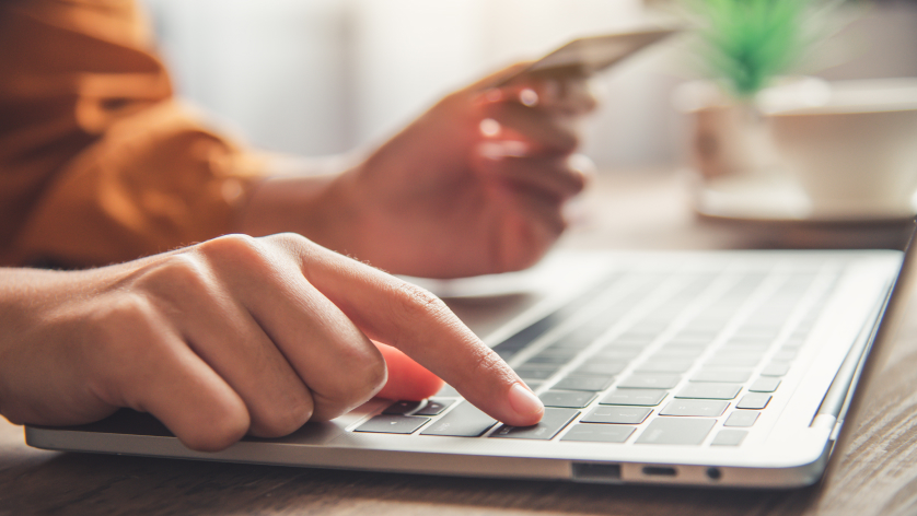 A person types on a laptop keyboard with one hand while holding a credit card in the other, suggesting online shopping, electronic payment, or managing an escrow bank account. A blurred cup and plant are visible in the background.