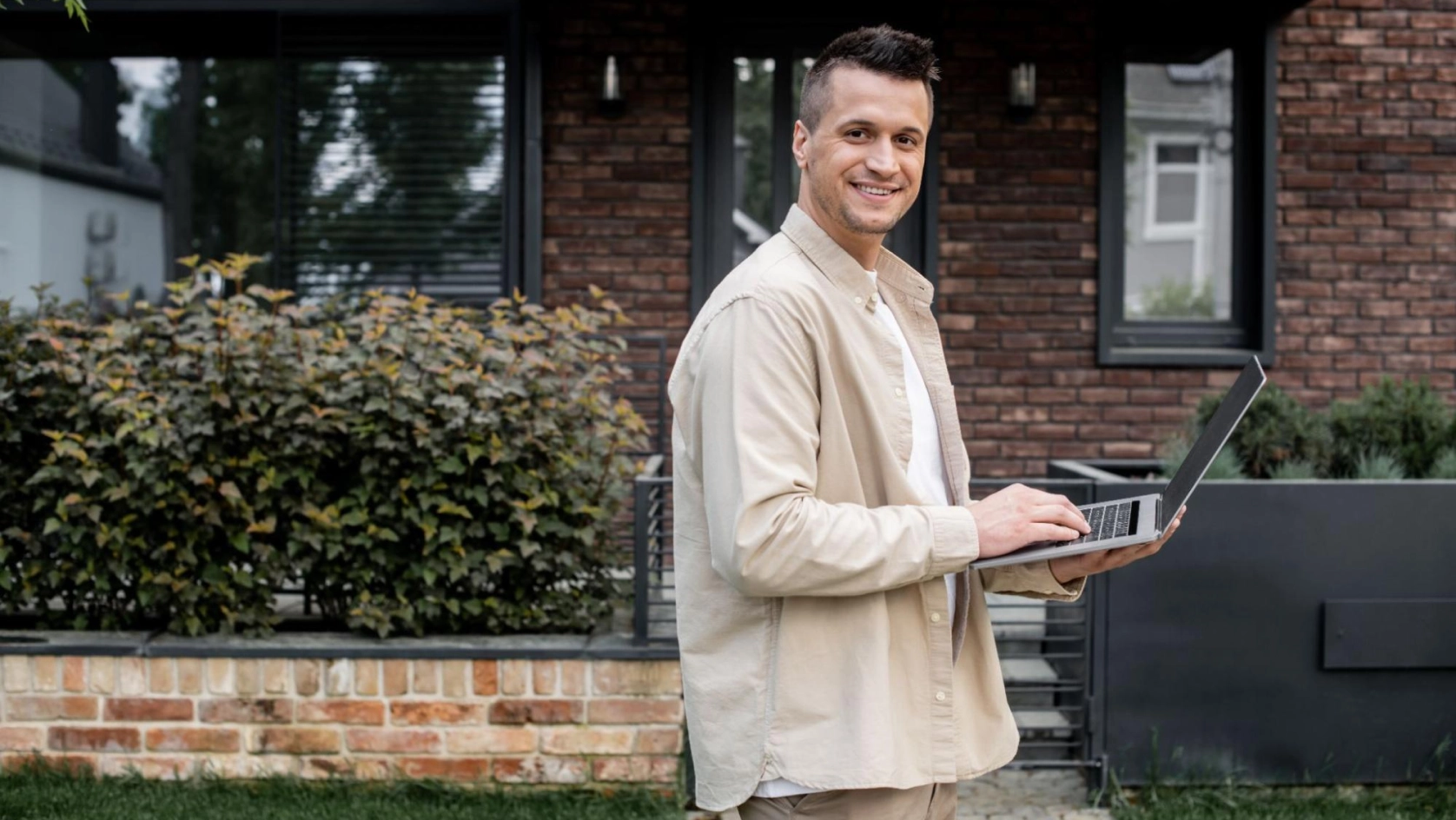 A man standing outside in front of a brick building, smiling while holding and typing on an open laptop. Shrubs and large windows are visible in the background.