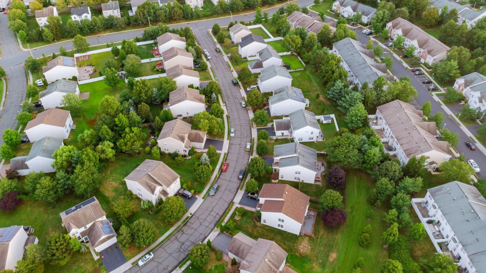 Aerial view of a suburban neighborhood with curved streets, single-family homes, and green lawns. Cars line the road, and houses with driveways hint at modern amenities like an AI bank account for real estate transactions.