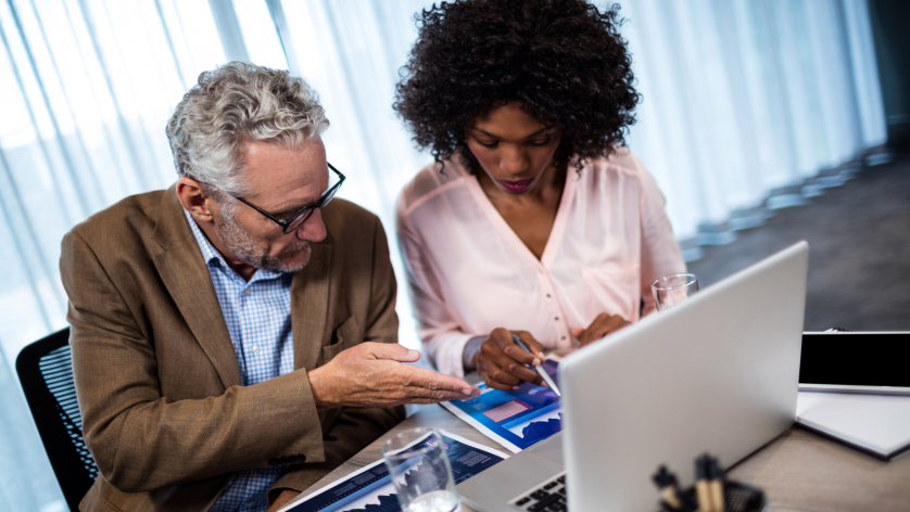 Two colleagues, a man in a brown blazer and a woman in a white blouse, discuss property management banking charts and graphs at a desk with a laptop and papers in a modern office setting.
