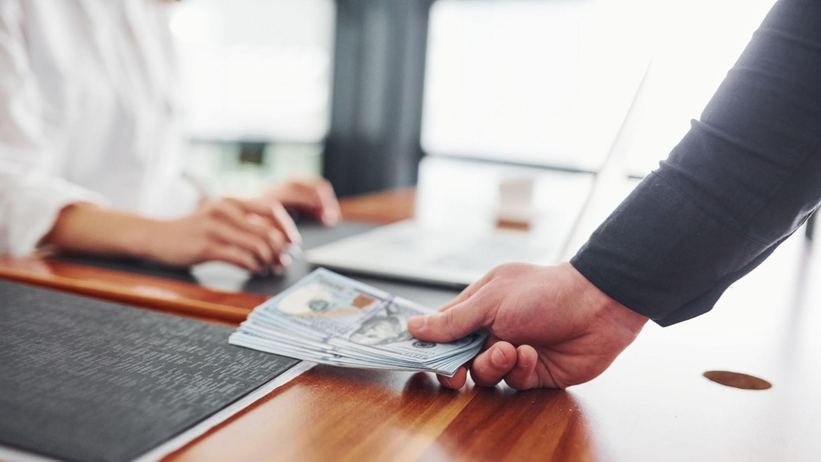 A person hands a stack of U.S. hundred-dollar bills to another individual sitting at a desk with a laptop, suggesting a financial or business transaction.