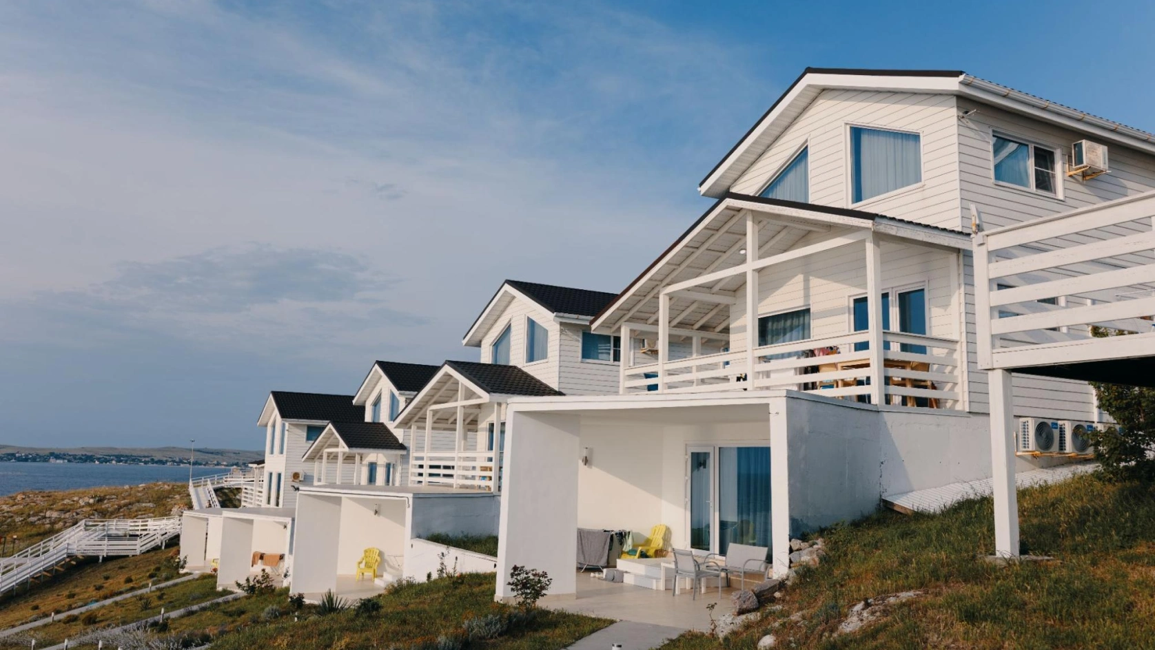 White seaside cottages with large windows and balconies overlook the ocean. Each house, ideal for an ai bank account for real estate investment, features outdoor seating and is built on a grassy hill under a partly cloudy sky.