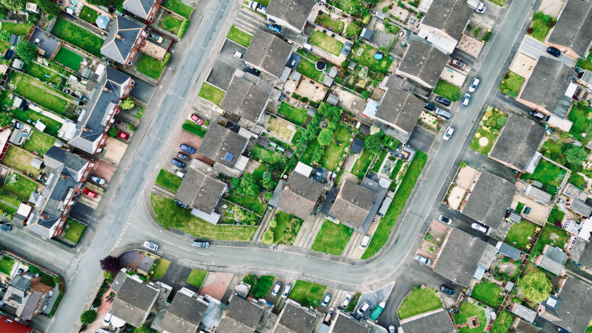 Aerial view of a suburban neighborhood with houses, neatly arranged gardens, streets, and parked cars. The roads form a curved intersection, and each house has a green yard.