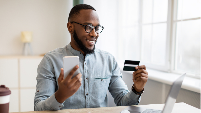 A smiling man wearing glasses holds a smartphone in one hand and a credit card in the other while sitting at a desk with a laptop, appearing to make an online purchase.