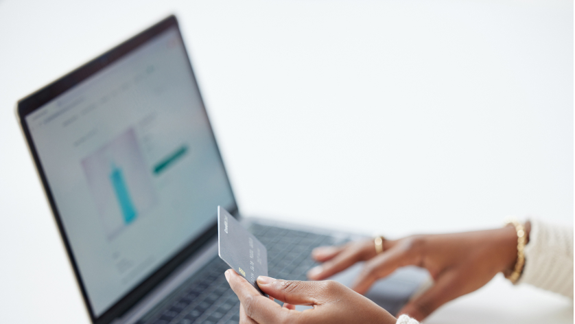 A person holds a credit card in one hand while using a laptop, appearing to make an online purchase. The laptop screen displays a shopping website with a product image.